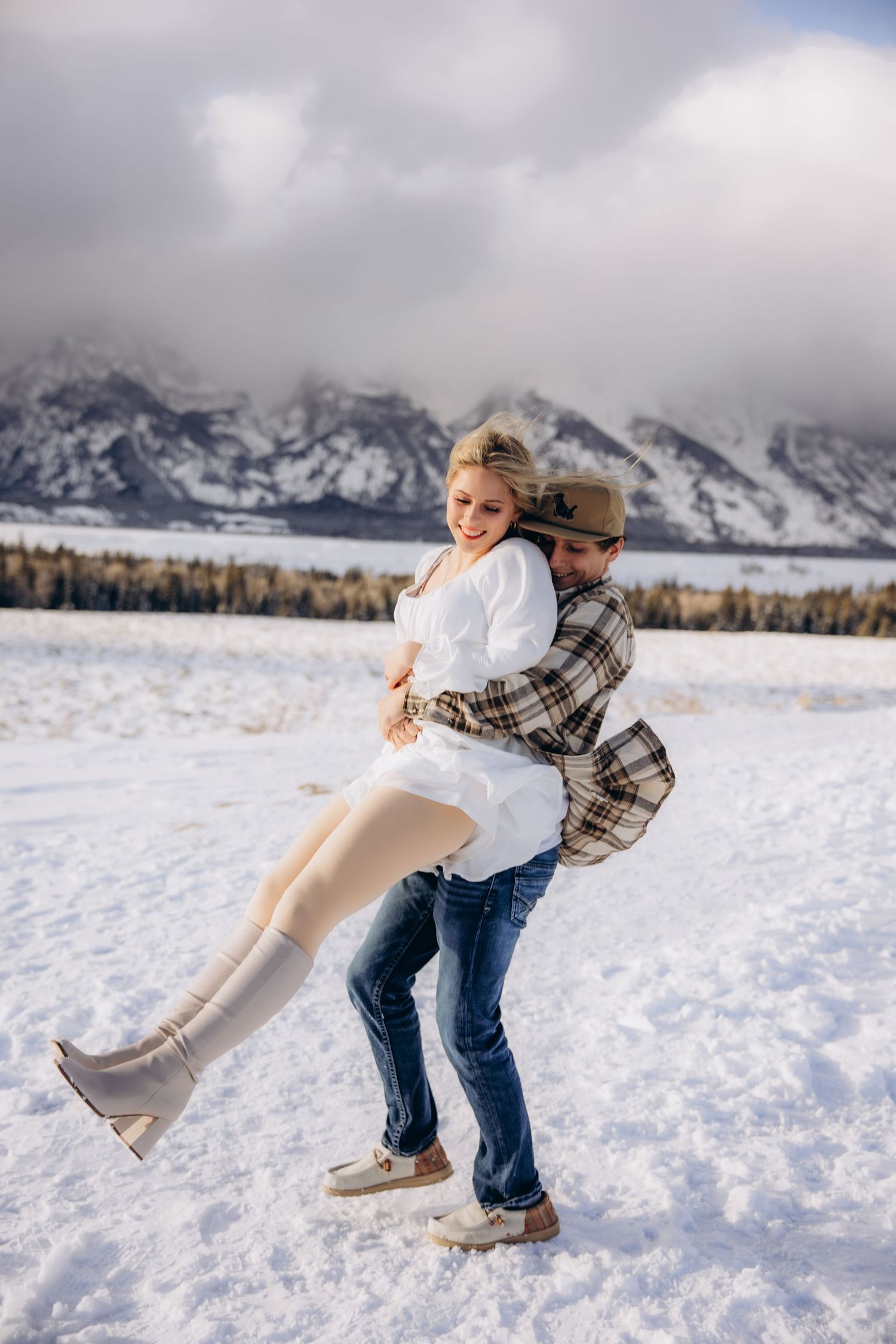 A playful winter scene of a couple embracing in the snow with majestic snow-capped mountains and forest in the background.