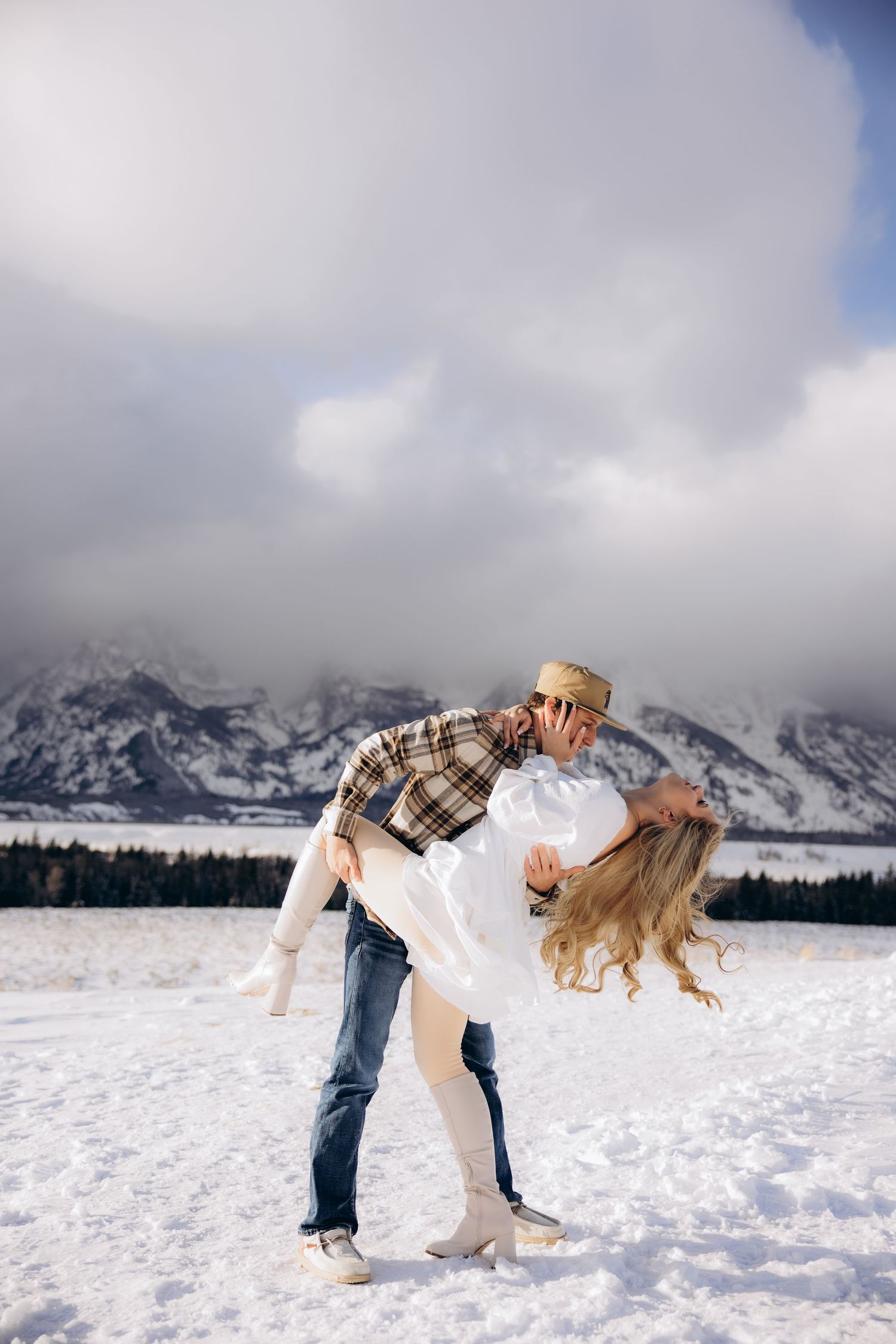 A romantic winter scene shows a couple dancing in the snow with snow-capped mountains and cloudy skies in the background.
