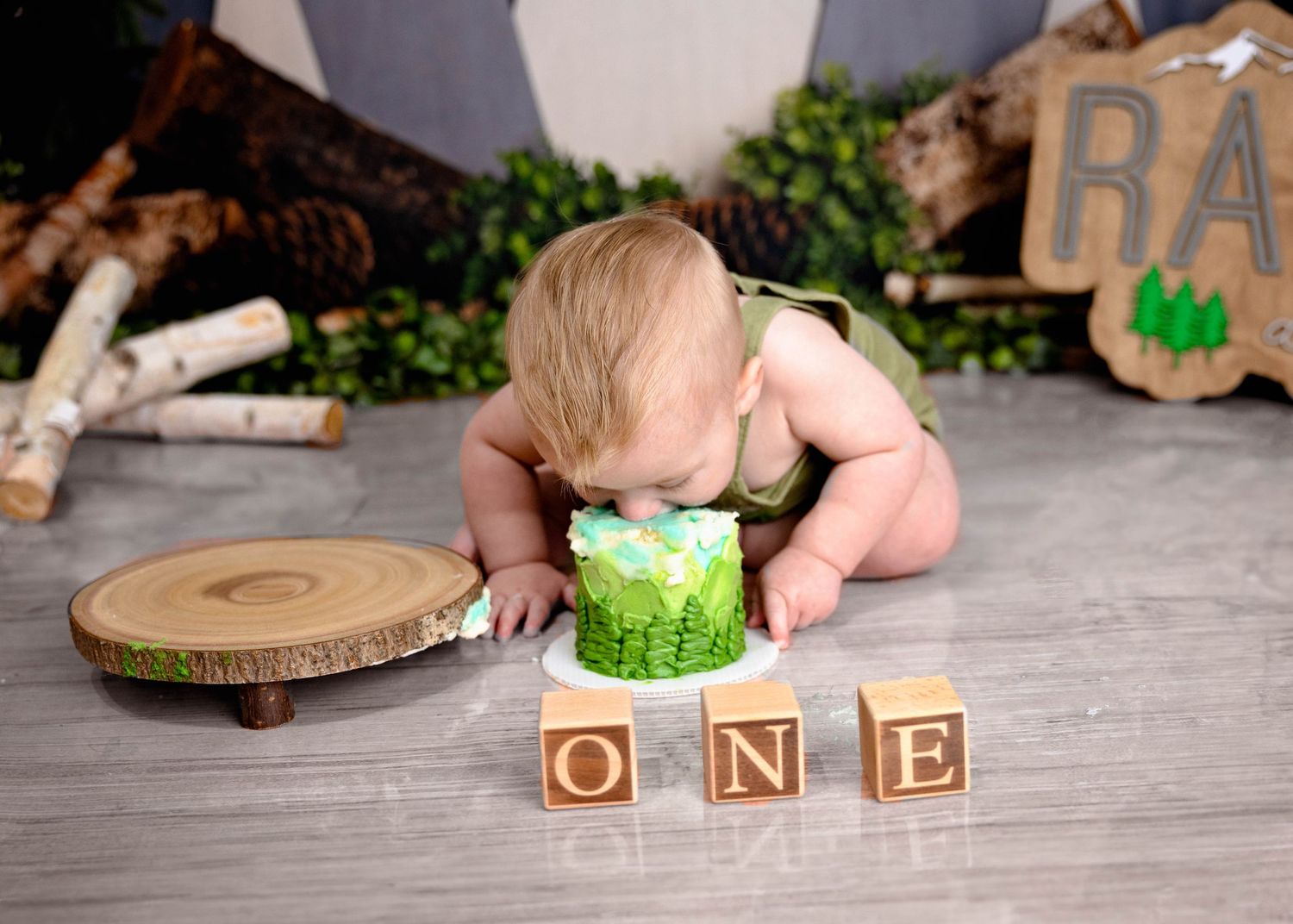A baby in a diaper leans forward to investigate a green smash cake with wooden 'ONE' blocks displayed in front. Cake Smash Carlsbad NM