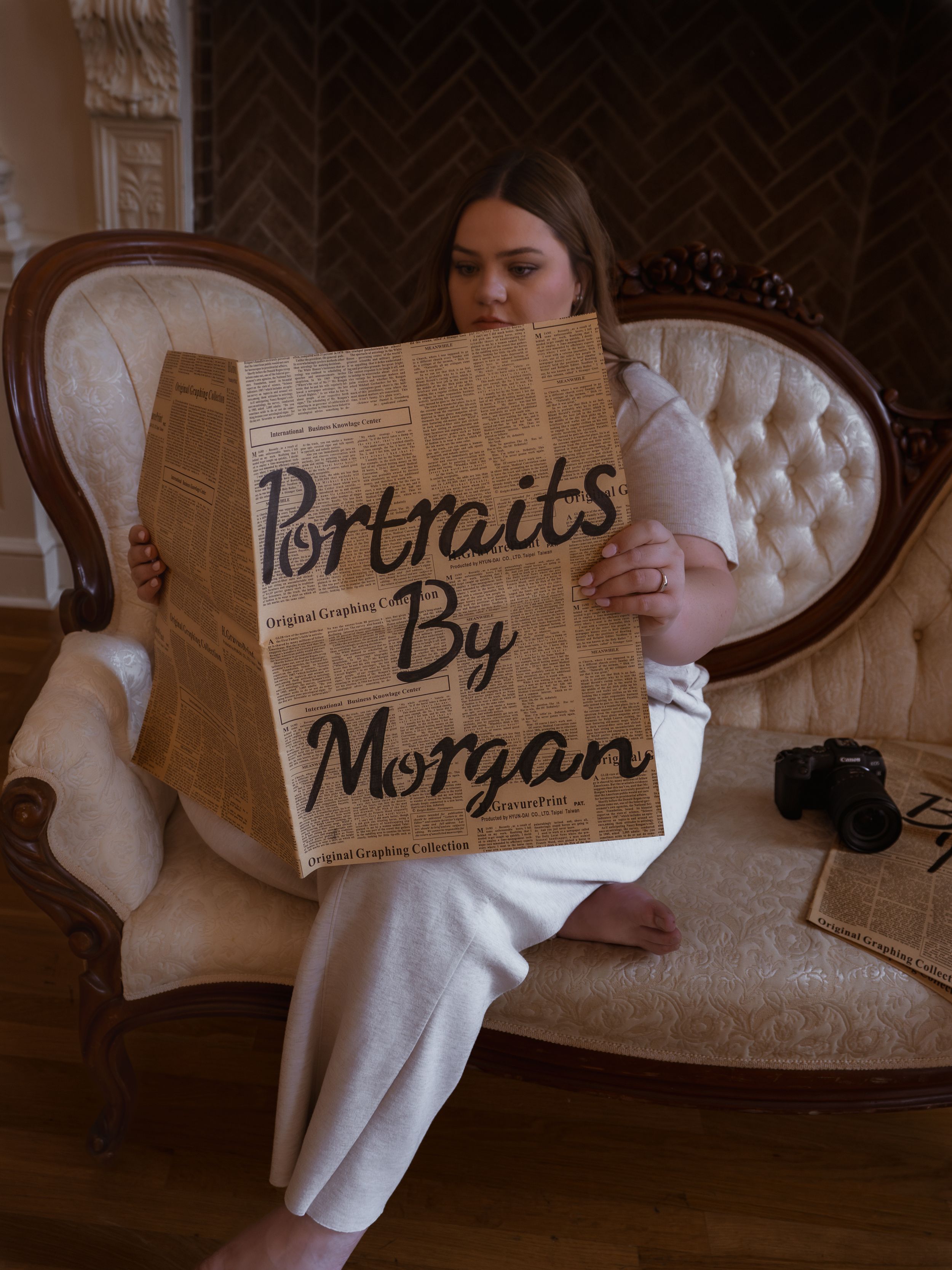 A person sits on an elegant tufted sofa holding a brown paper portfolio labeled "Portraits by Morgan" with a camera nearby.