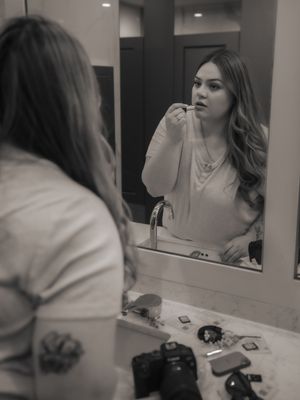 A person applies makeup while looking in a bathroom mirror with a camera and cosmetics visible on the counter.