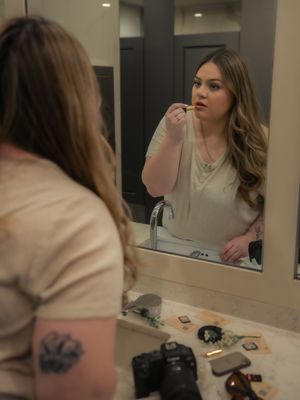 A person applies makeup in front of a bathroom mirror with cosmetic products visible on the counter.