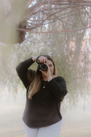 Photographer in black clothes takes pictures in a misty forest setting with soft lighting and atmospheric fog.