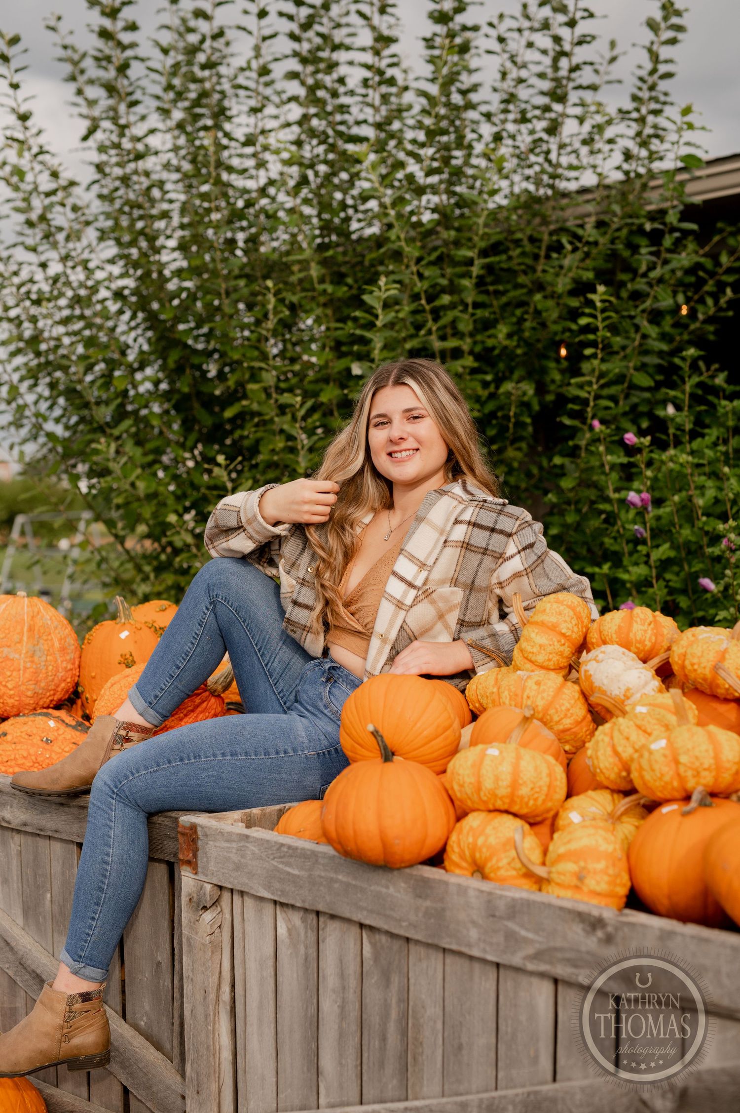 Someone in jeans and plaid shirt relaxes among orange pumpkins on rustic wooden display.