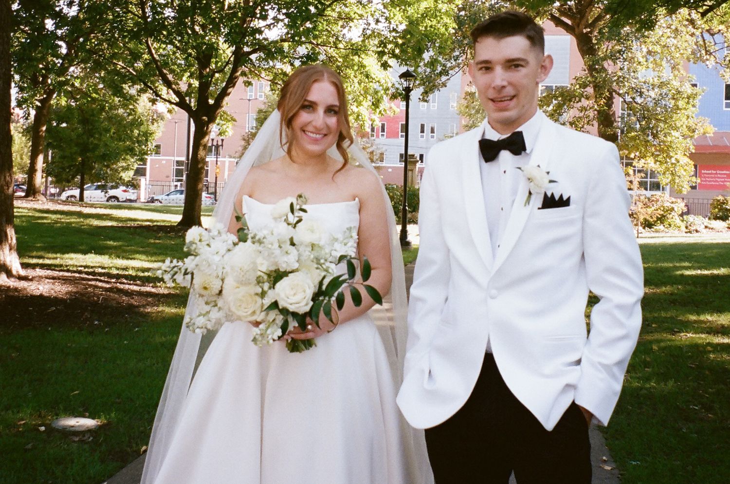 A bride in white gown with bouquet poses with groom in white tuxedo outdoors.