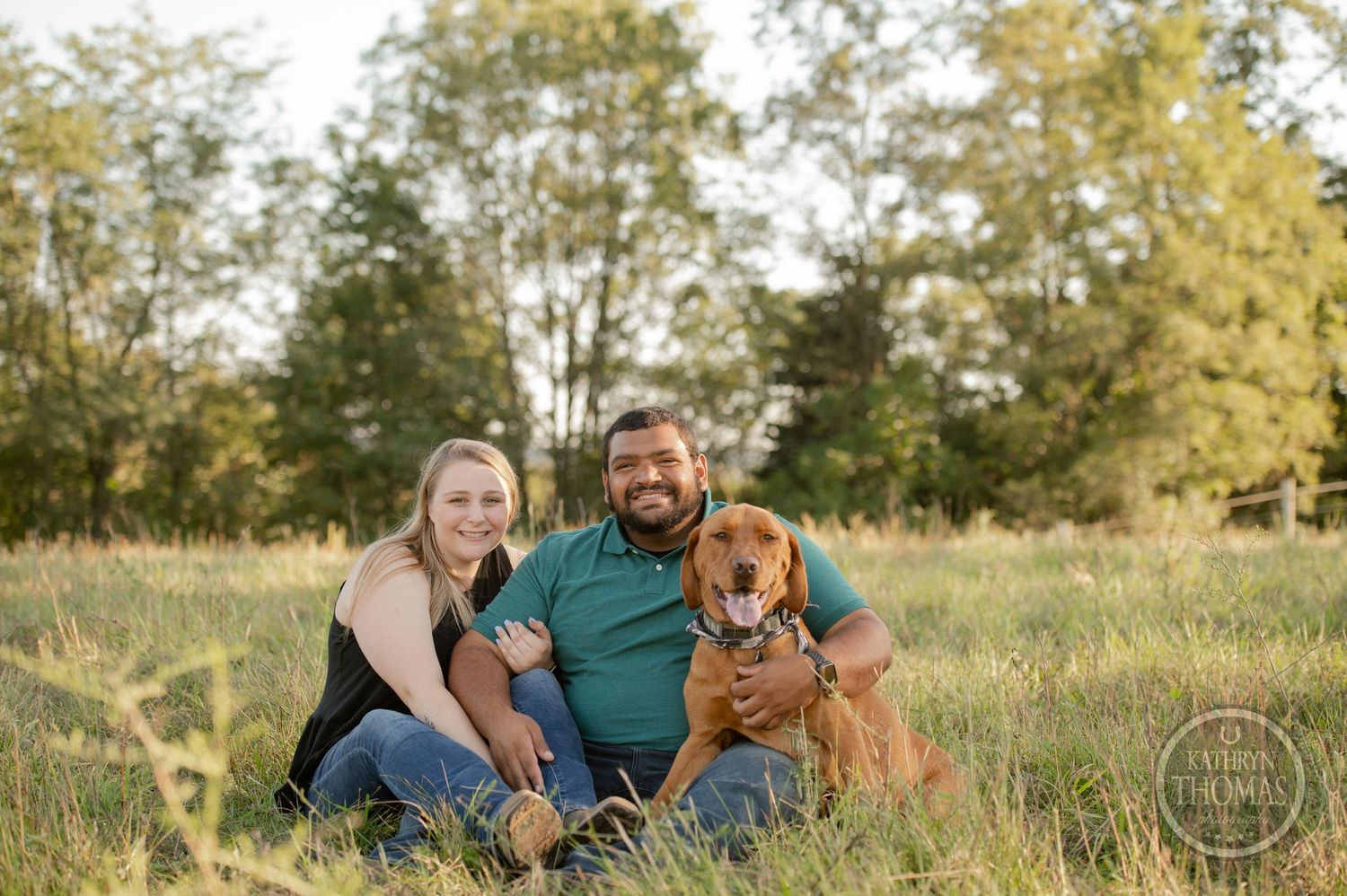 A couple sits in a sunny field with their brown dog during a photo session.