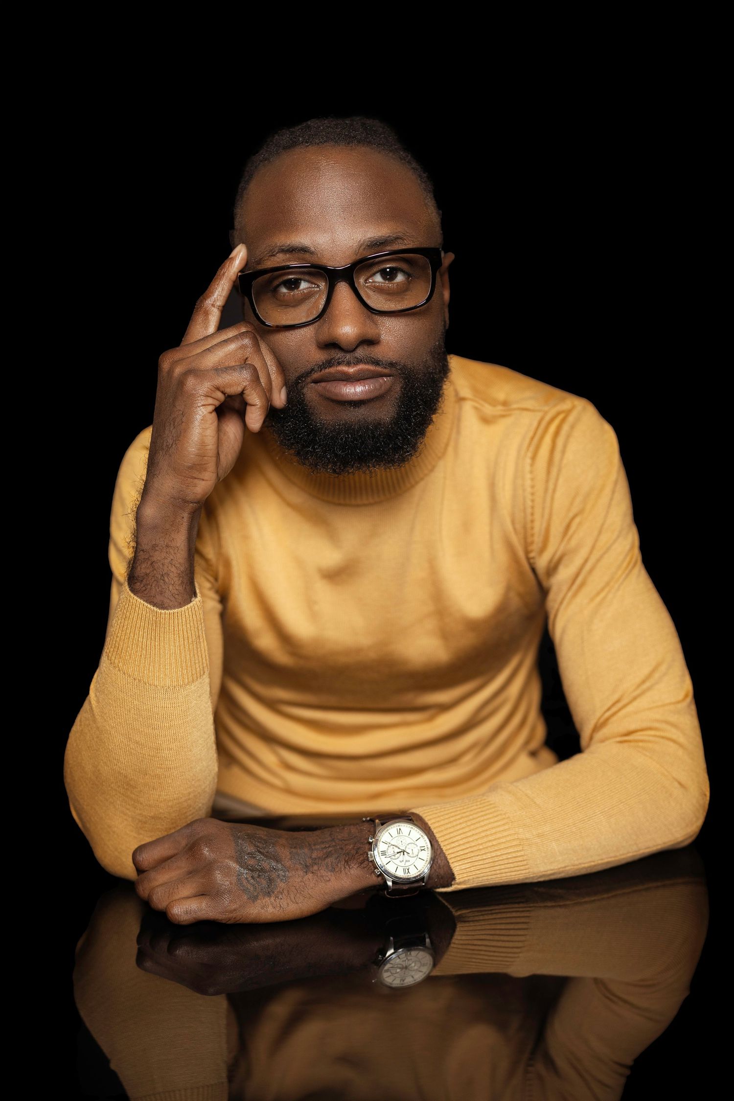 Professional portrait of a person in a yellow sweater wearing glasses and a watch, with hand thoughtfully placed near temple.