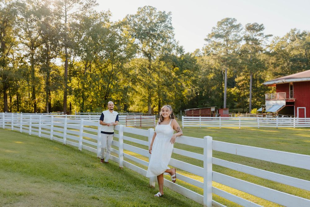 A couple walks and poses along a white picket fence on a sunny afternoon.