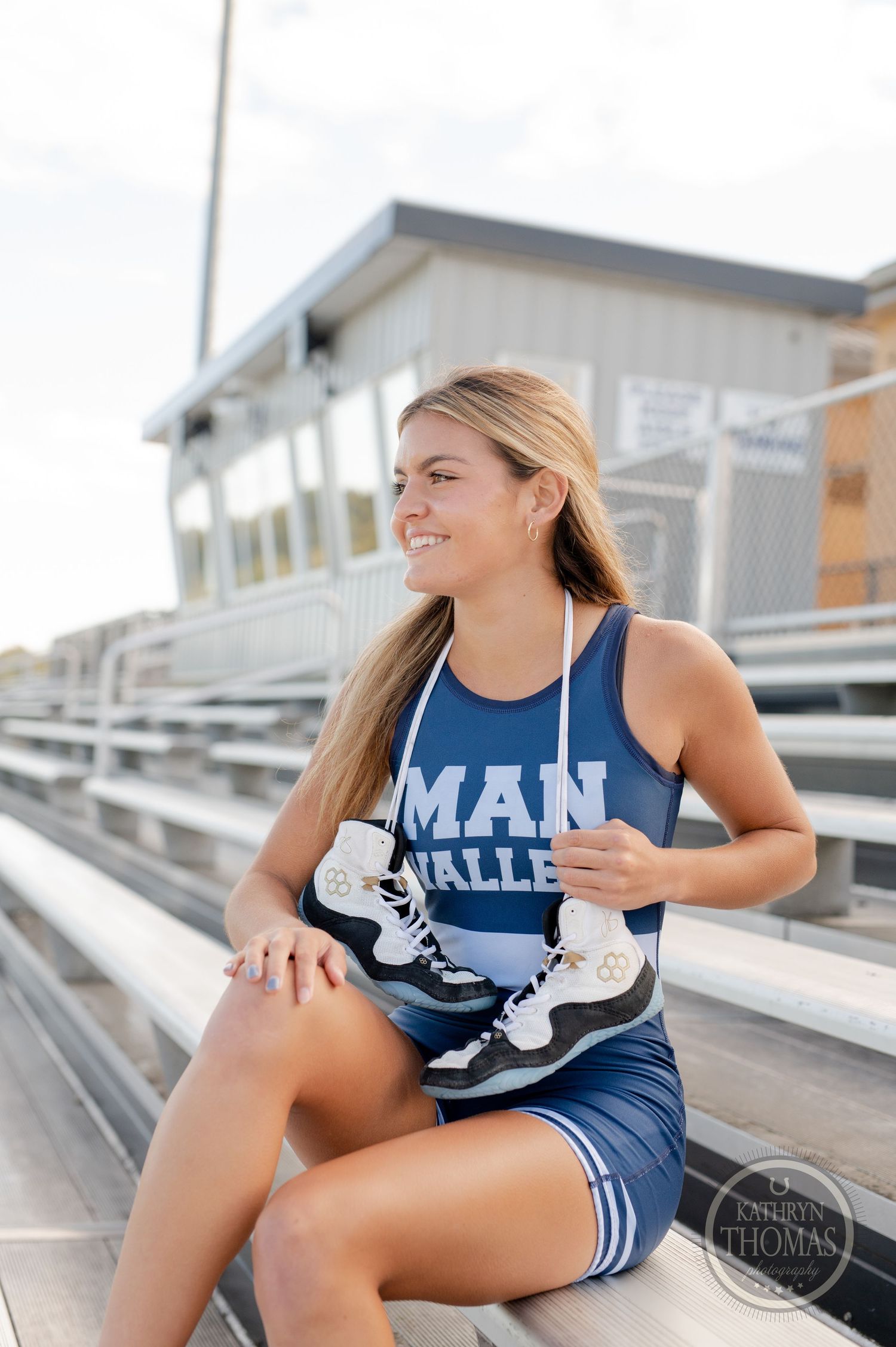 Athlete in blue uniform sits casually on stadium bleachers holding athletic shoes.