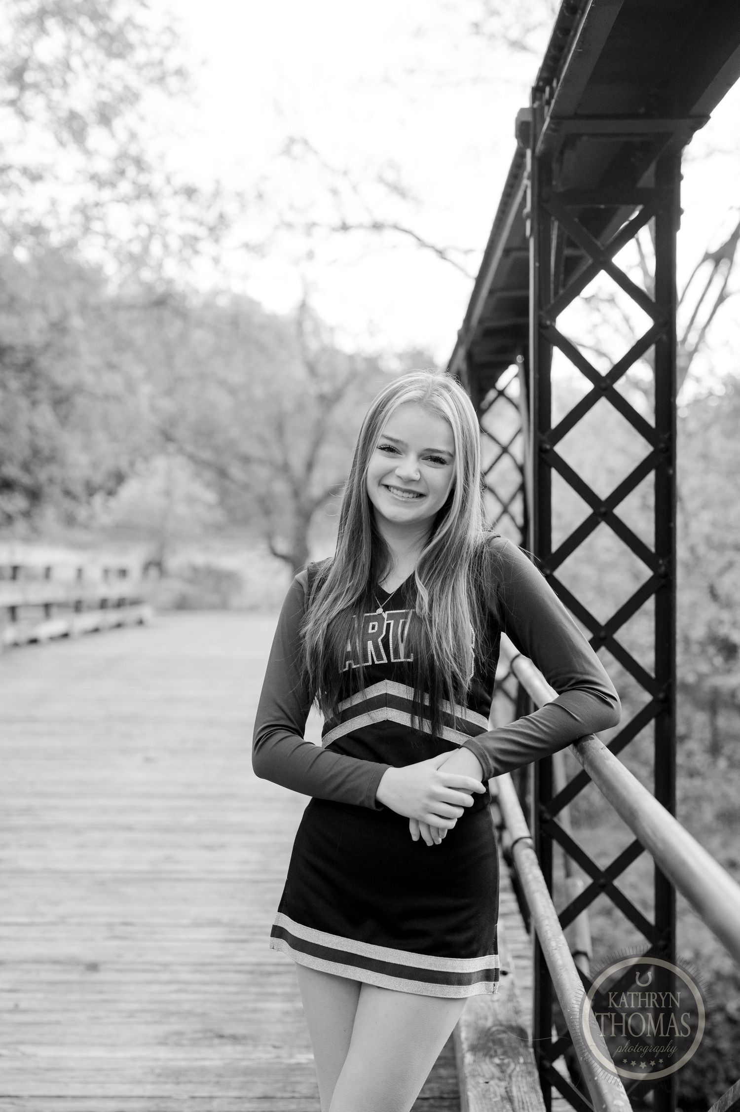 Black and white portrait of cheerleader posing on wooden bridge with metallic support beams.
