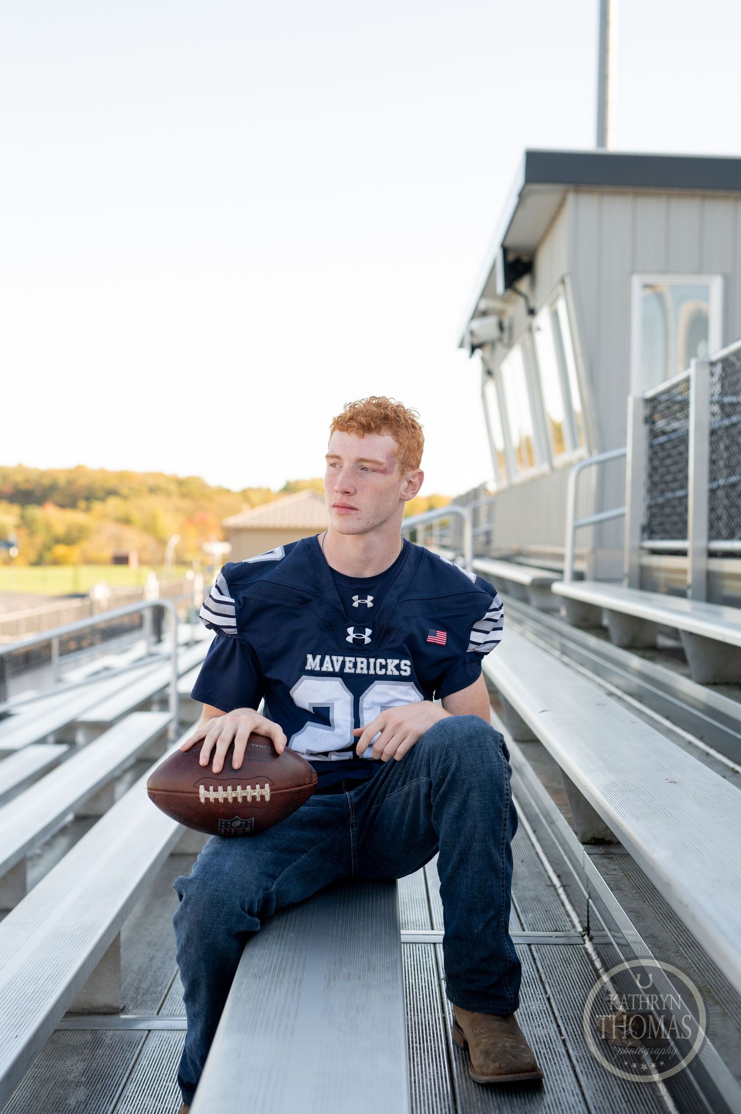 Football player in Mavericks jersey holds ball while sitting on stadium bleachers.