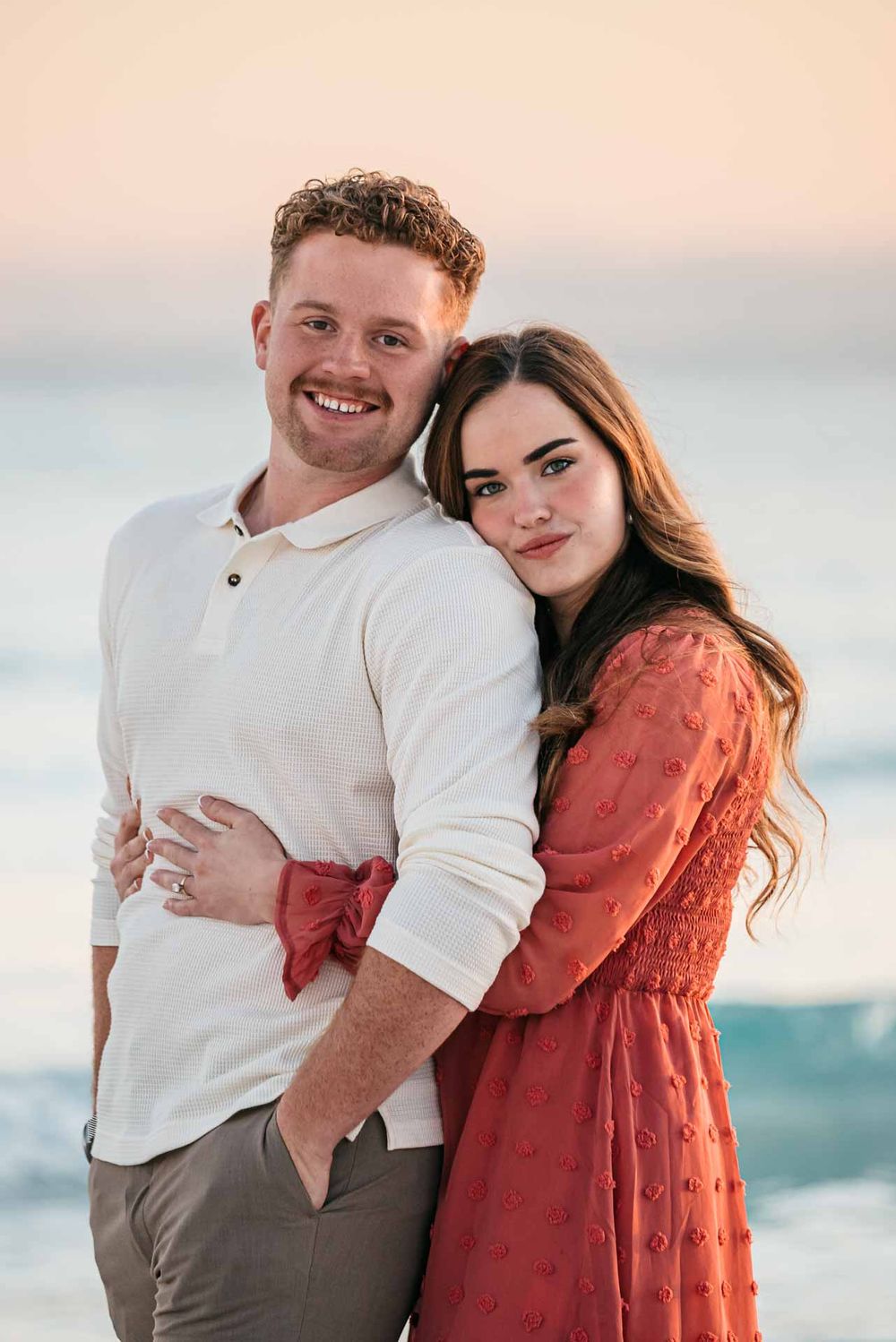 A romantic couple embraces on the beach at sunset wearing white and coral colored outfits.