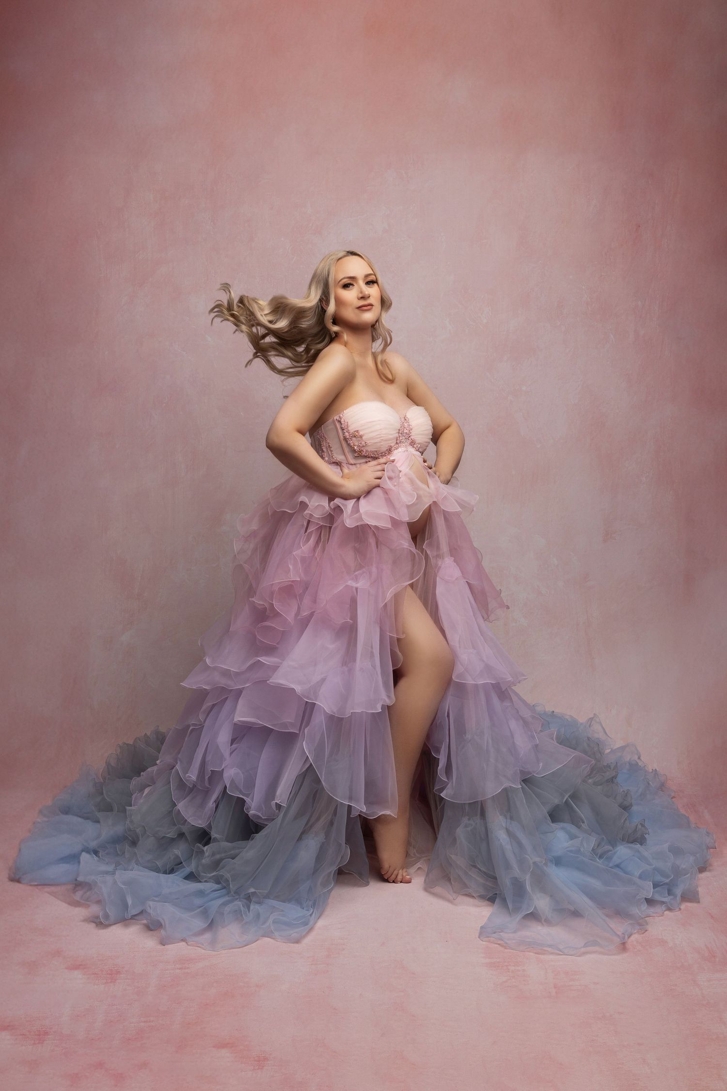 A glamorous portrait in a flowing pink to blue ombre tulle gown with ruffled layers against a pink backdrop.