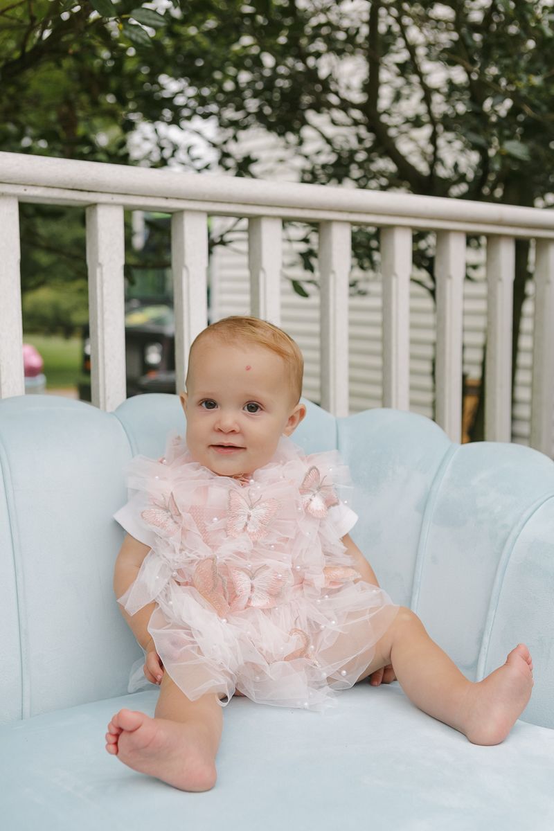 Birthday girl sitting on a soft blue sofa wearing a pink dress and smiling, captured by NYC family photographer Paula O
