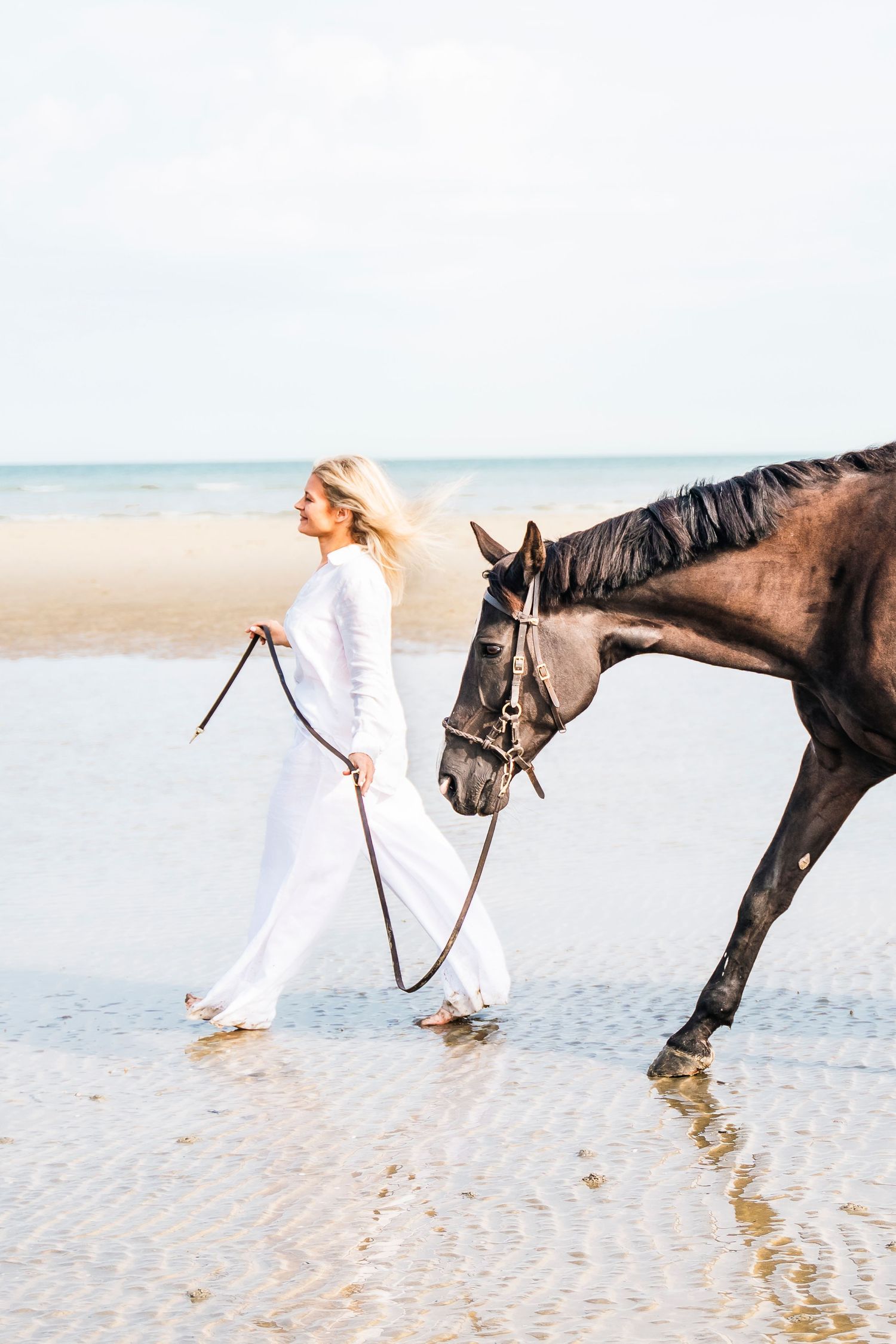 A person in white walks with a dark horse along the shoreline.