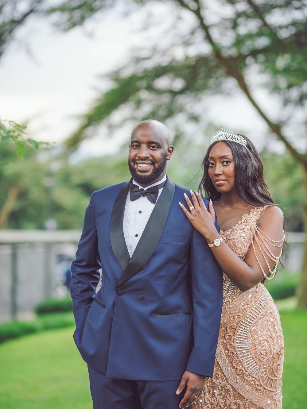 Elegant couple in formal wear posing on manicured lawn with trees in background.