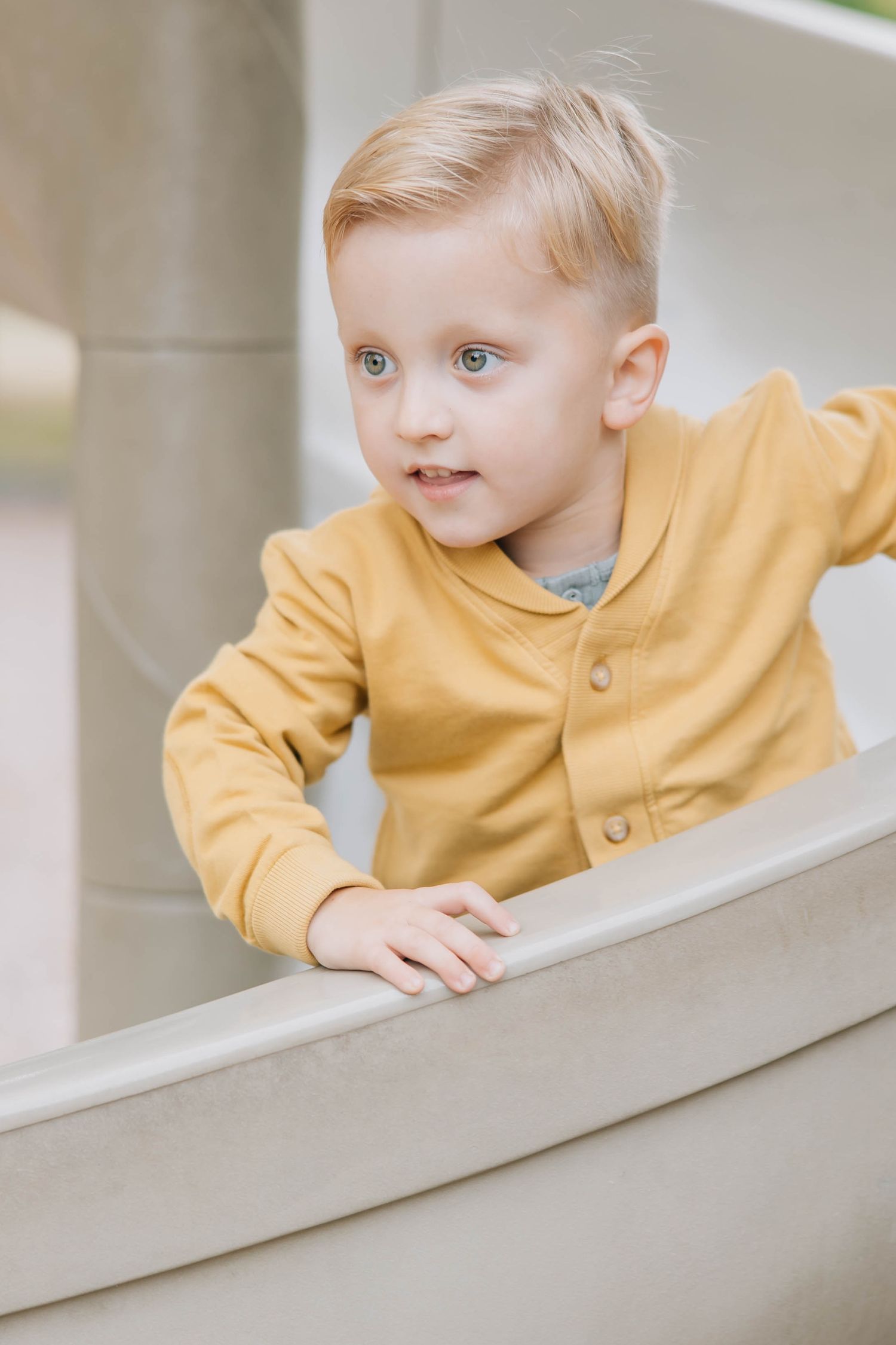 Cute blonde toddler boy in yellow shirt looking over a ledge during outdoor photo session.