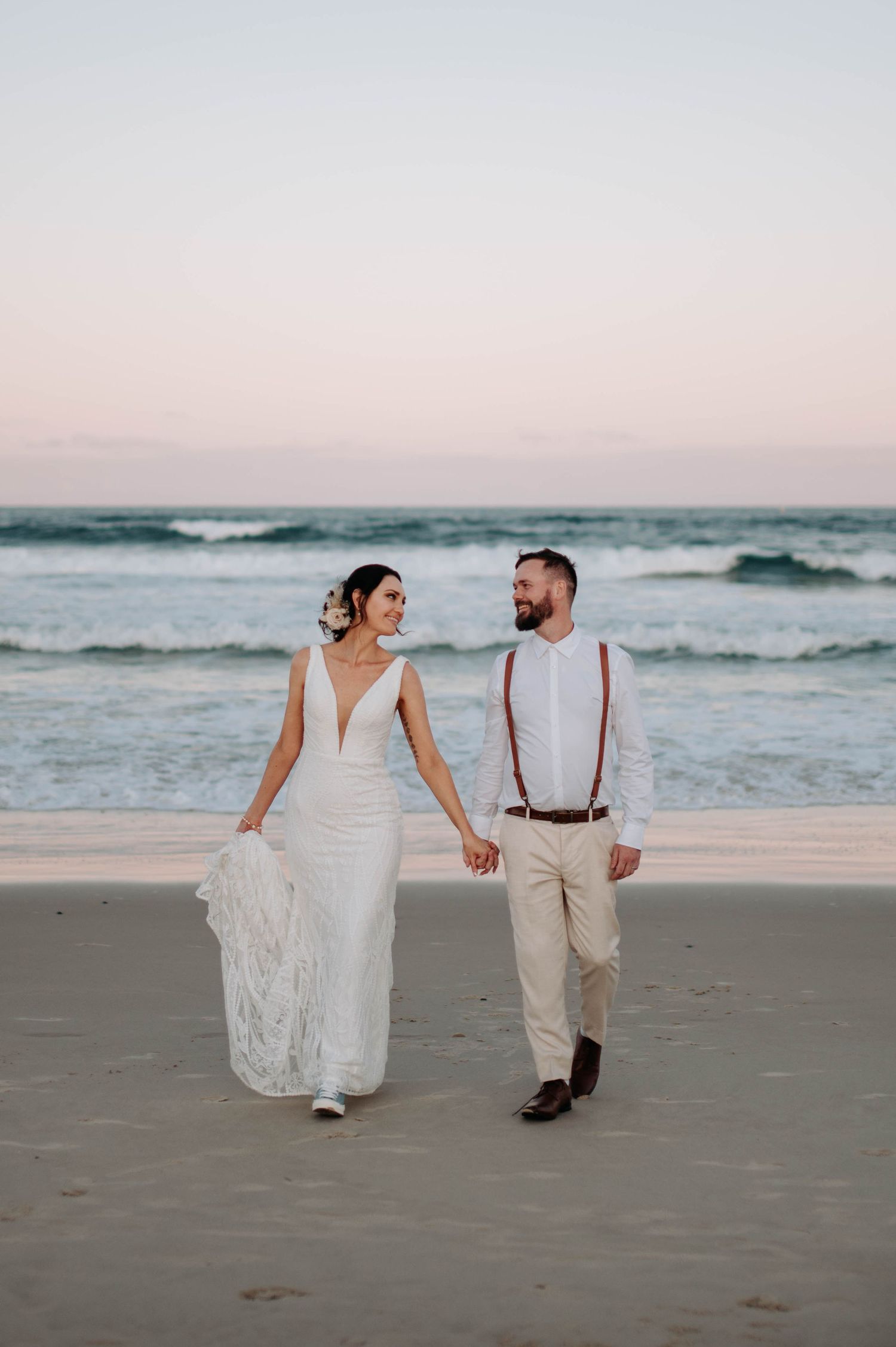 Couple walking hand in hand along beach shore at dusk in wedding attire.