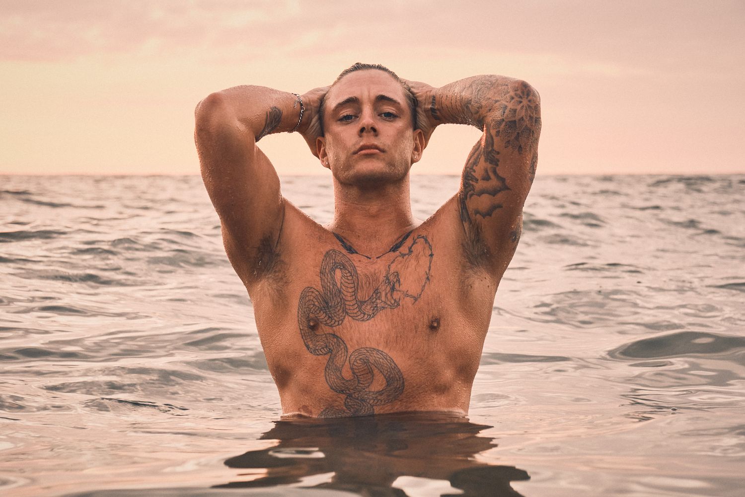 Tattooed male model standing in ocean at sunset, hands on head, exuding confidence with dramatic lighting