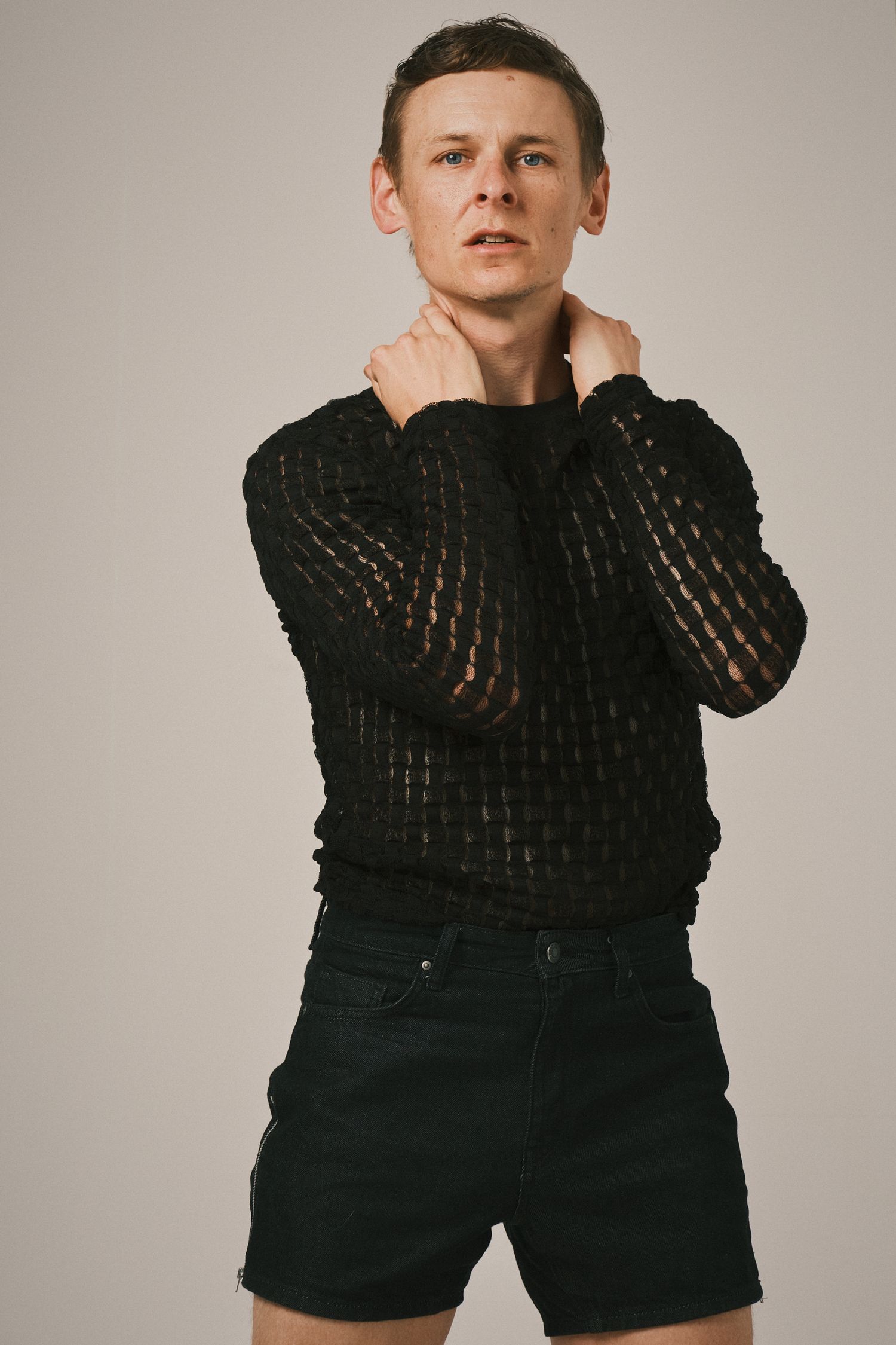 Male model in black mesh top and denim shorts, posing with hands on neck, showcasing a contemporary, edgy look