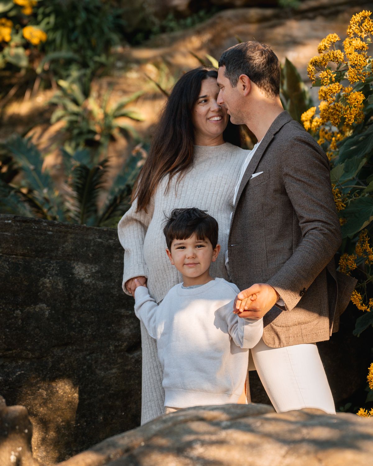 parents-looking-at-eachother-while-their-son-smiles-into-the-camera-during-a-family-photoshoot-among-nice-yellow-flowers