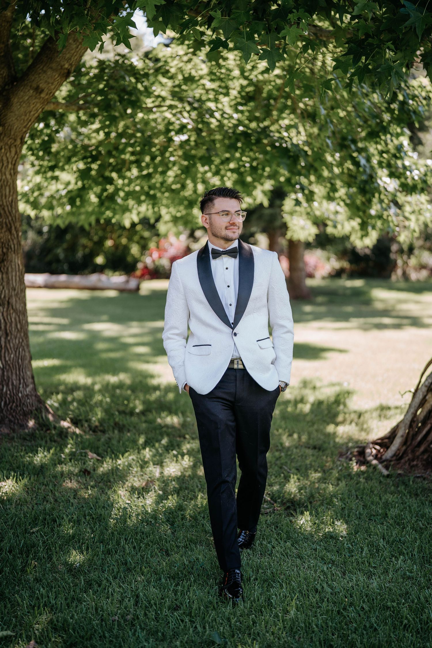 A groom in a white tuxedo jacket and black bow tie poses playfully in a sunny garden setting with lush green trees.