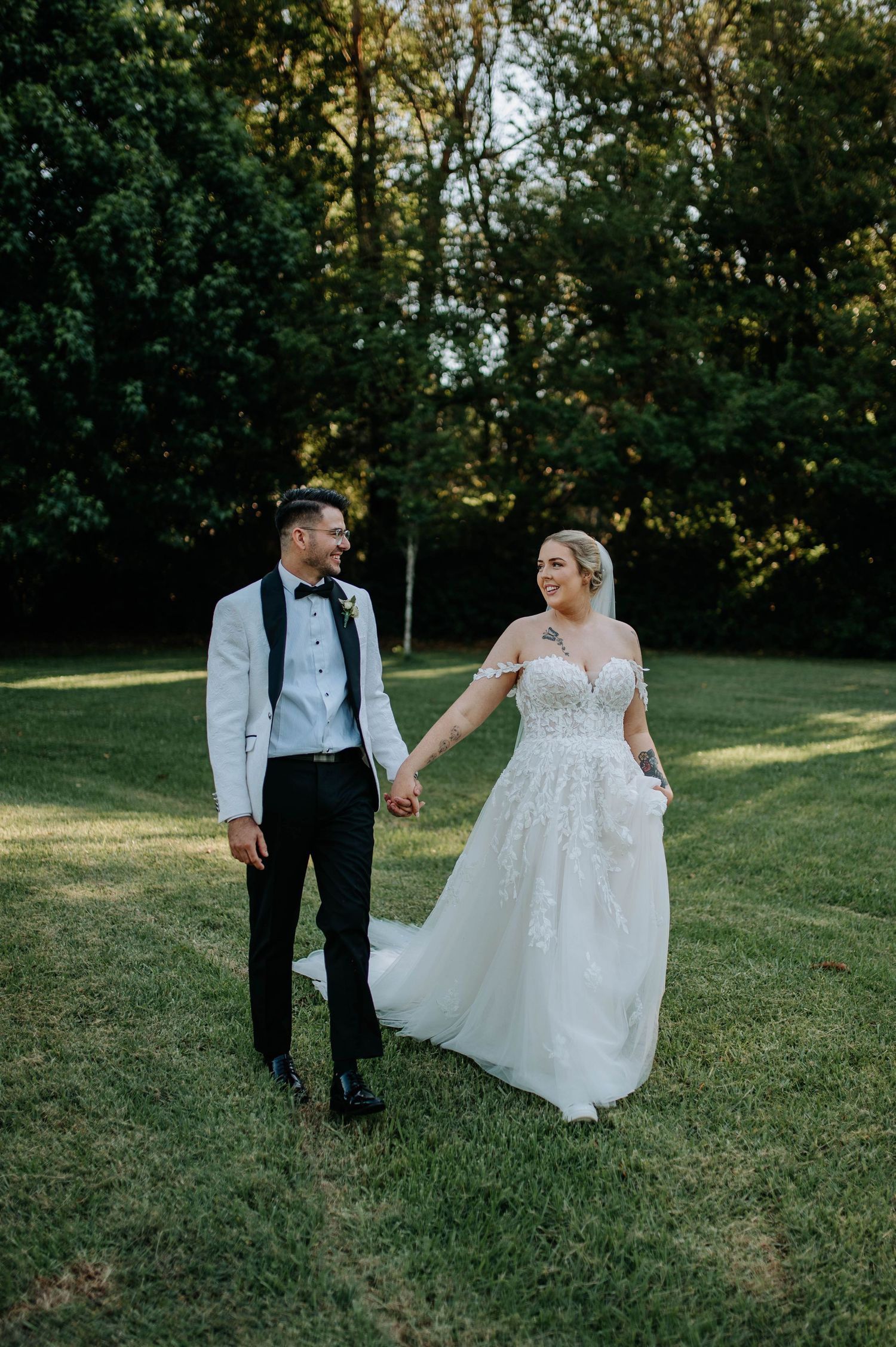 A couple in wedding attire walks together on a grassy hill during golden hour.