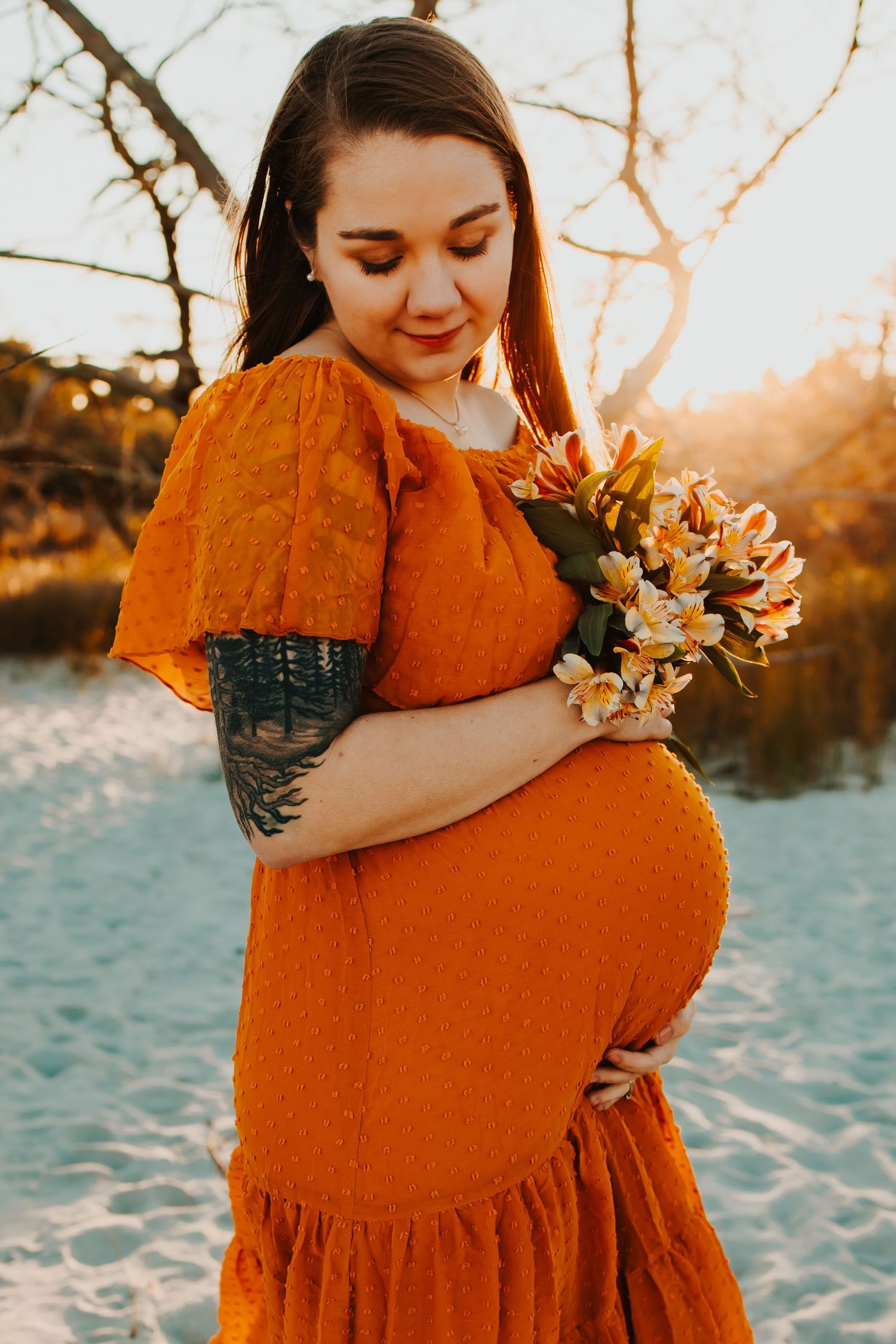 Maternity photos of woman in orange dress holding flowers against sunset at beach.