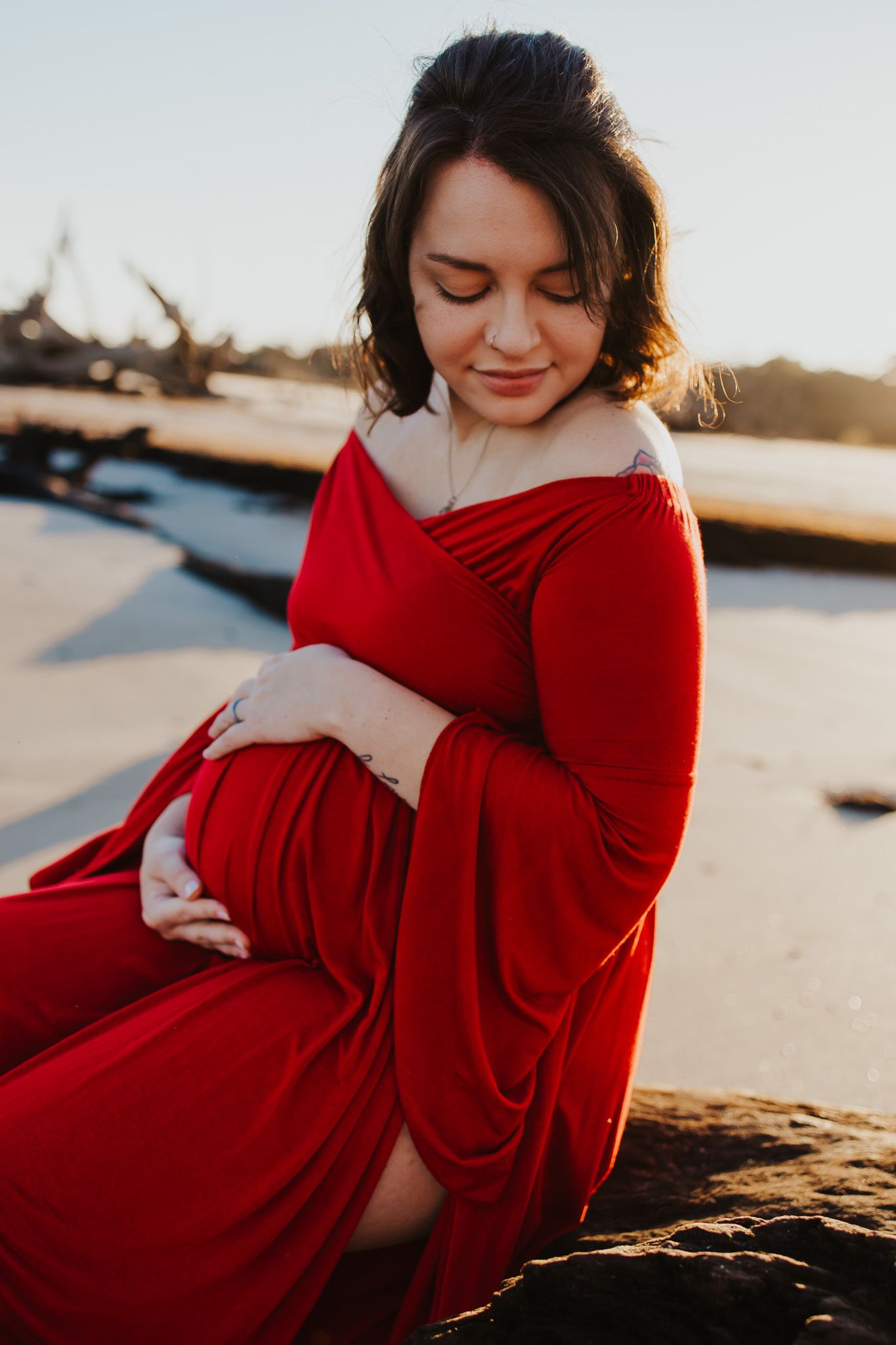 Expectant mother in red dress poses during sunset maternity photoshoot.