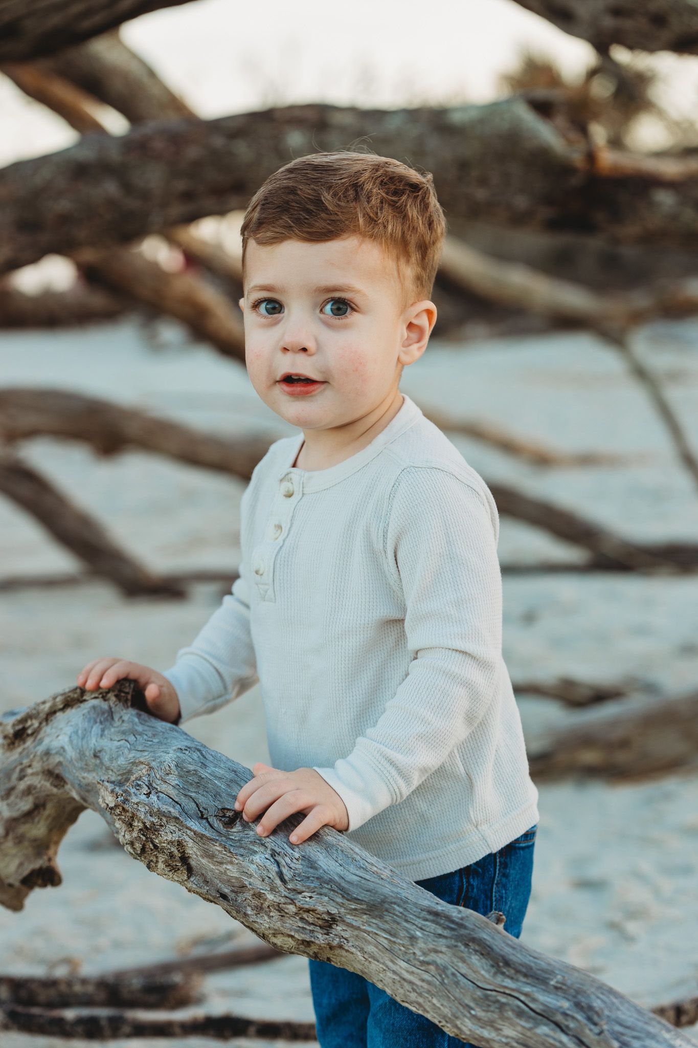 Young child in white henley shirt and jeans explores driftwood on a beach during sunset.