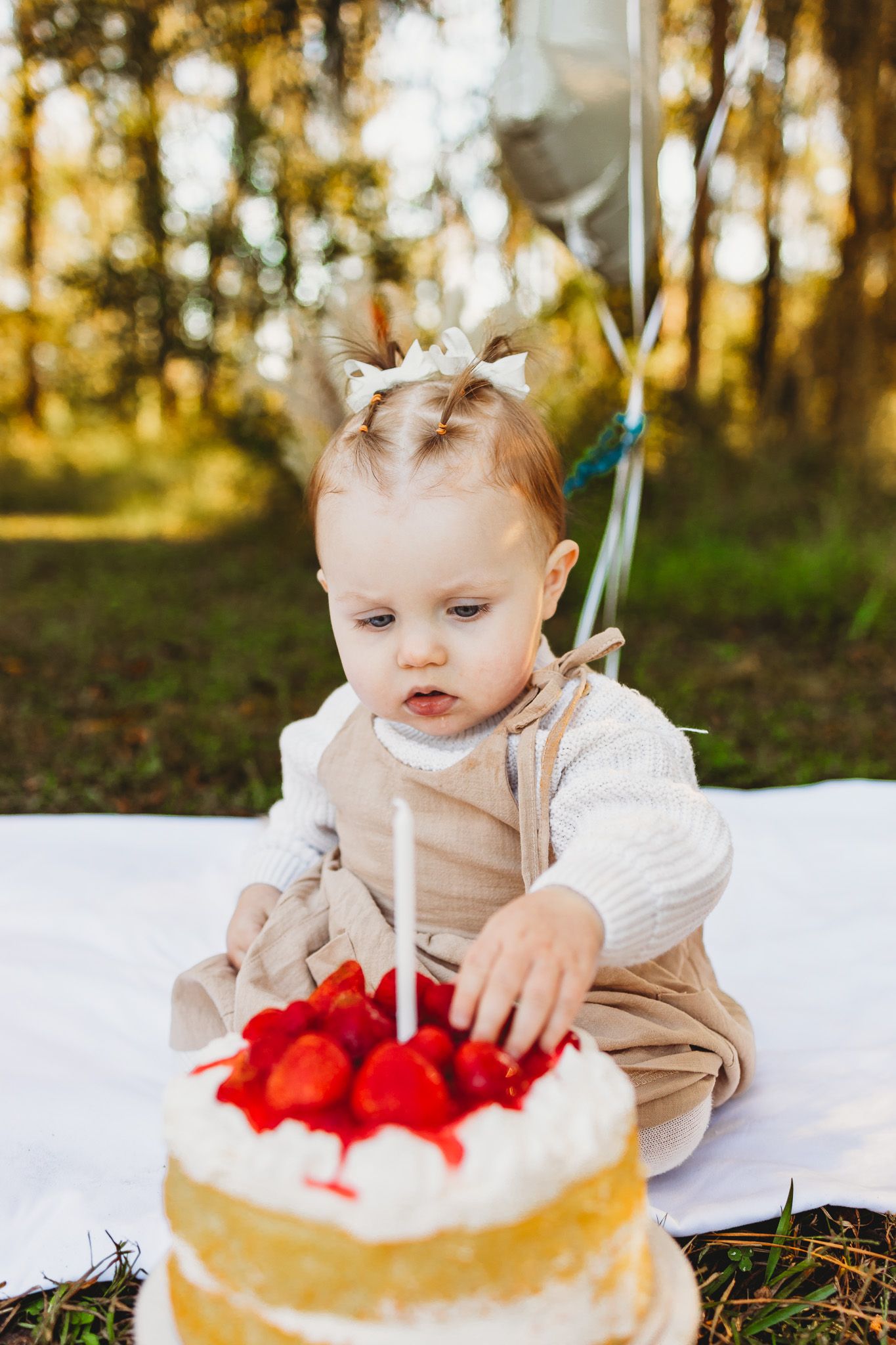Baby in beige outfit reaches for a birthday cake topped with red berries and candles on a white blanket outdoors.