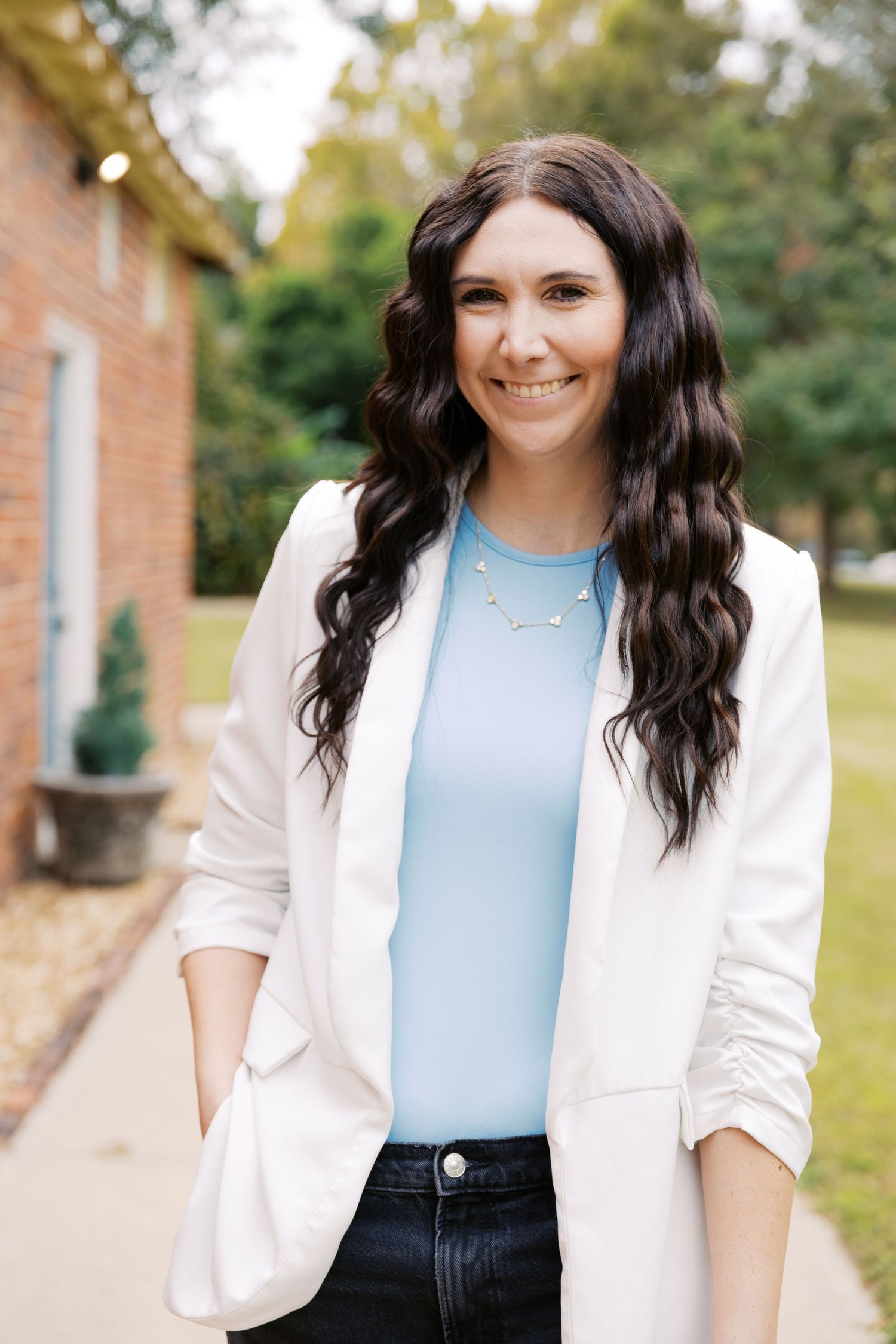 Woman in white blazer and blue top poses outdoors for professional photos.