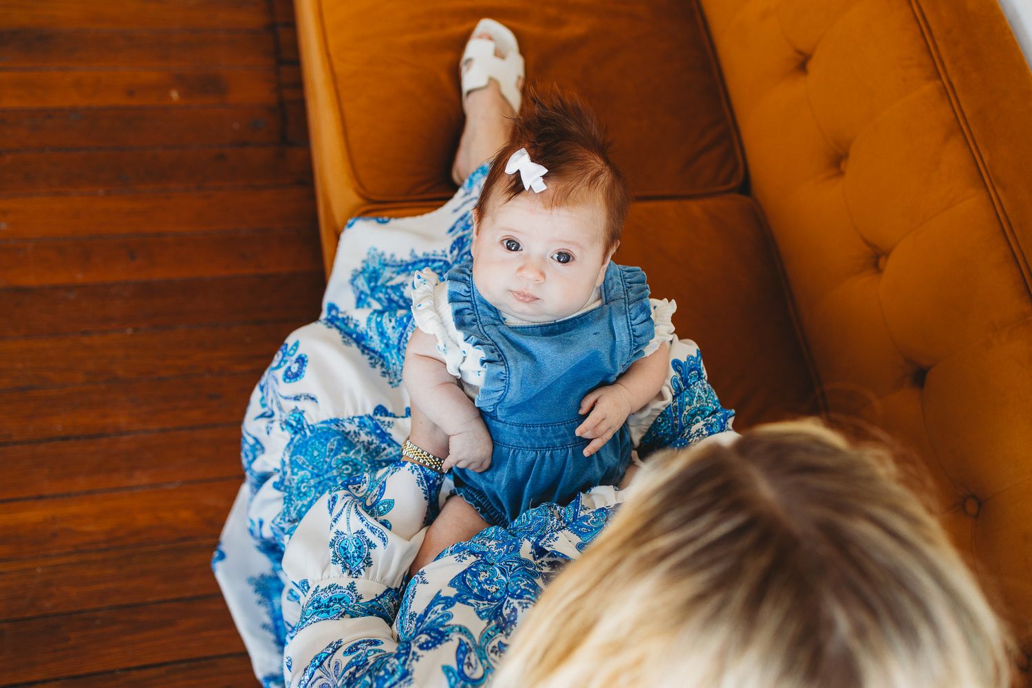 Baby in blue floral outfit sits on brown leather couch during indoor photoshoot.