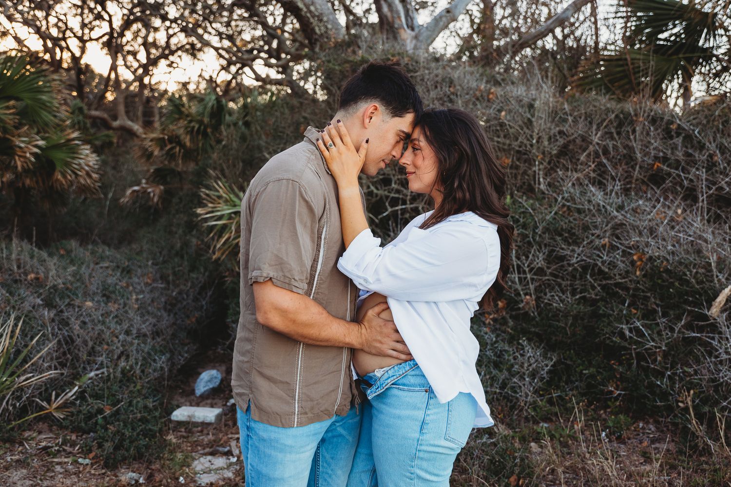 A couple embraces in front of bare tree branches during sunset in an outdoor setting.
