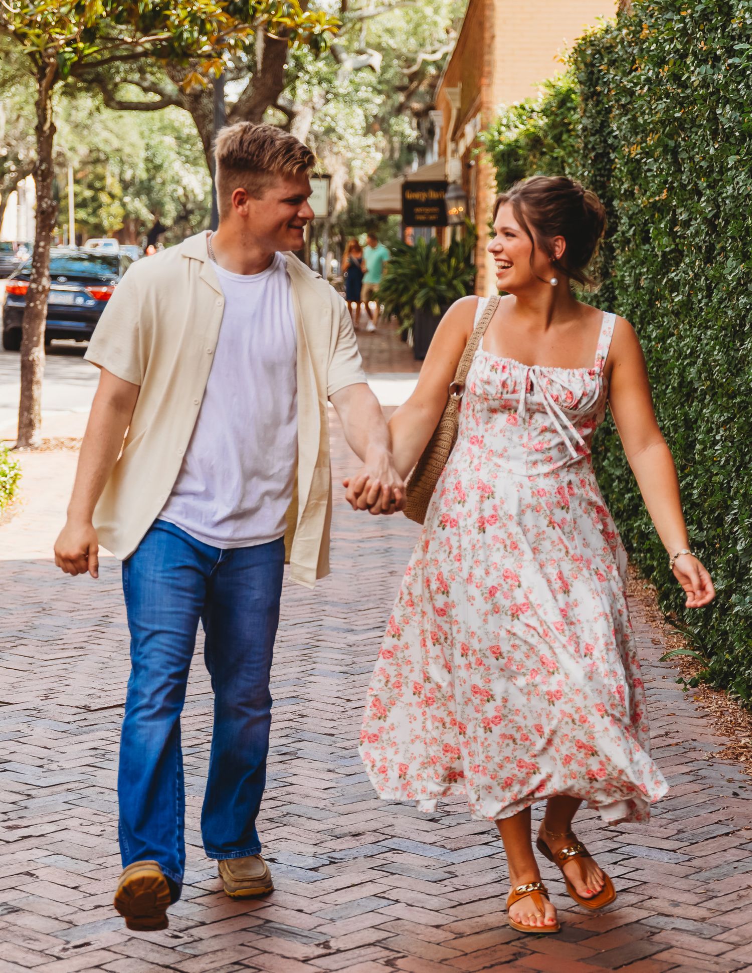 A couple strolls hand in hand down a charming brick street, with her wearing a floral dress and him in casual attire.