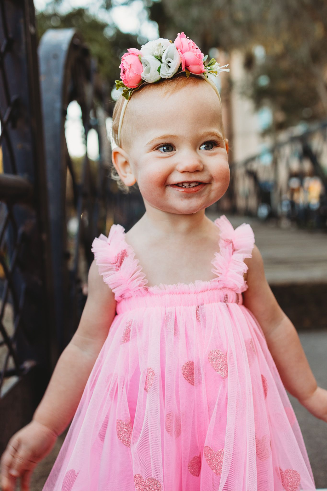A baby wearing a pink tulle dress and floral headband smiles joyfully in an outdoor setting.