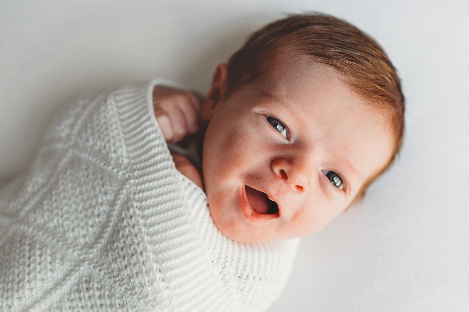 A baby wearing a white knit sweater lies on a light colored surface while making different facial expressions.