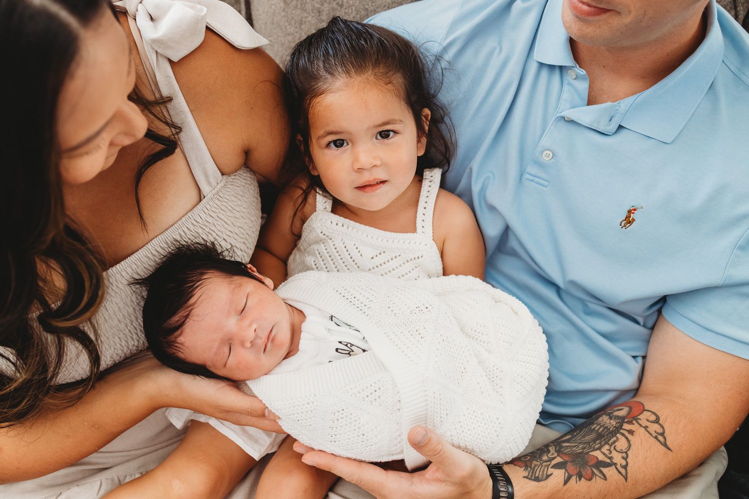 Family sitting together on couch with newborn baby and young child in white dress.