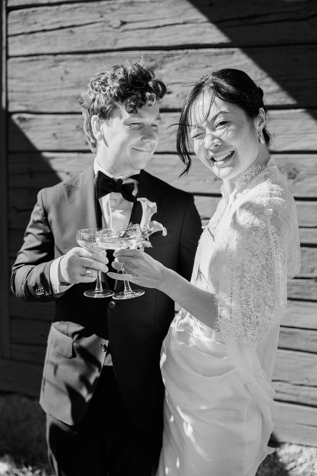 A couple in wedding attire share a toast with champagne glasses on a wooden deck in black and white.