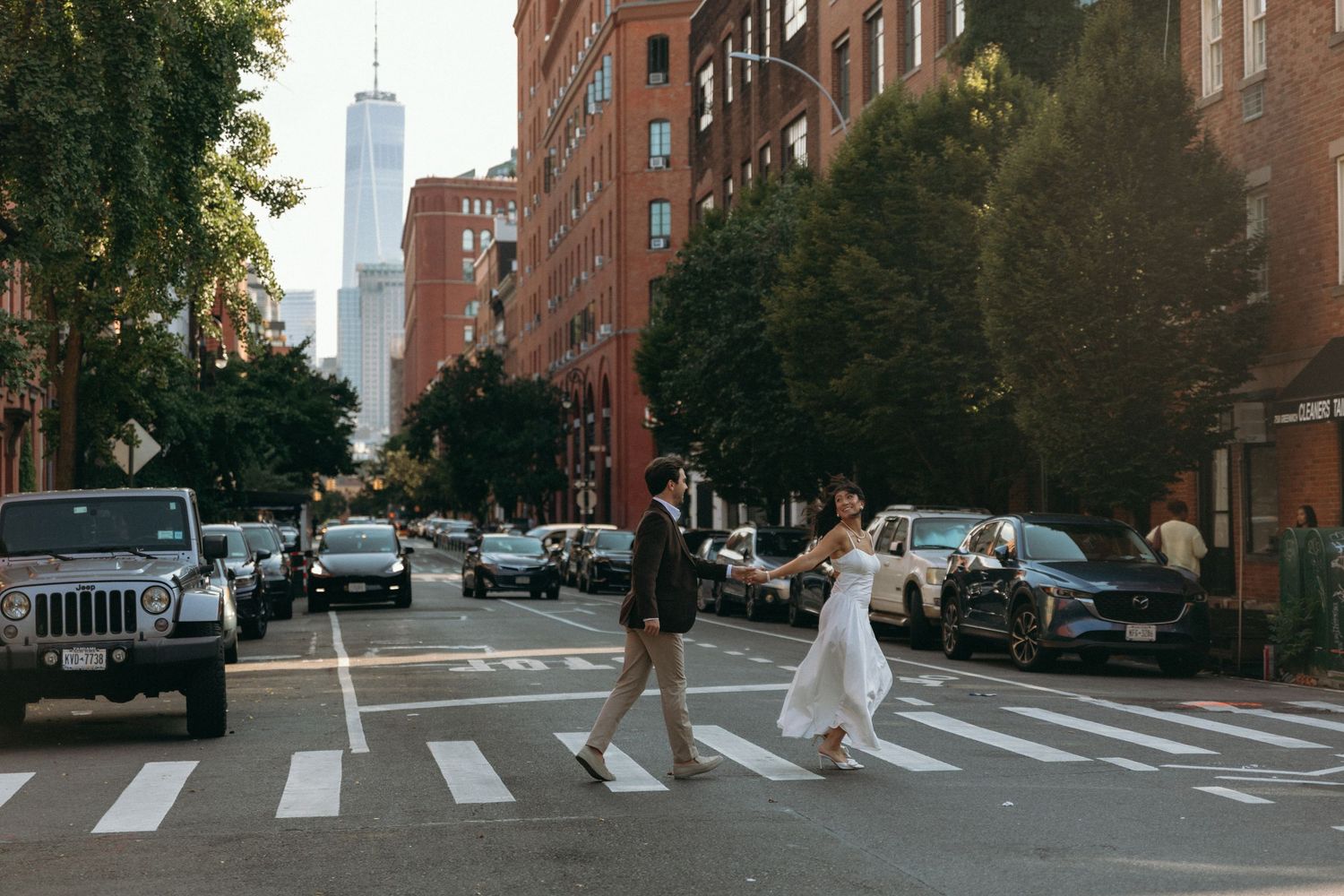 Two people cross a street in New York City with the Freedom Tower visible in the background on a sunny day.