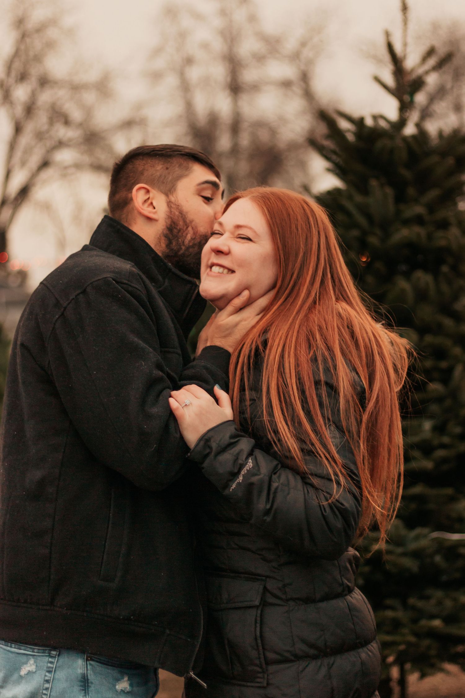 Couple embracing outdoors in winter, man kissing woman's cheek, she smiles | Westminster Colorado Proposal Photographer