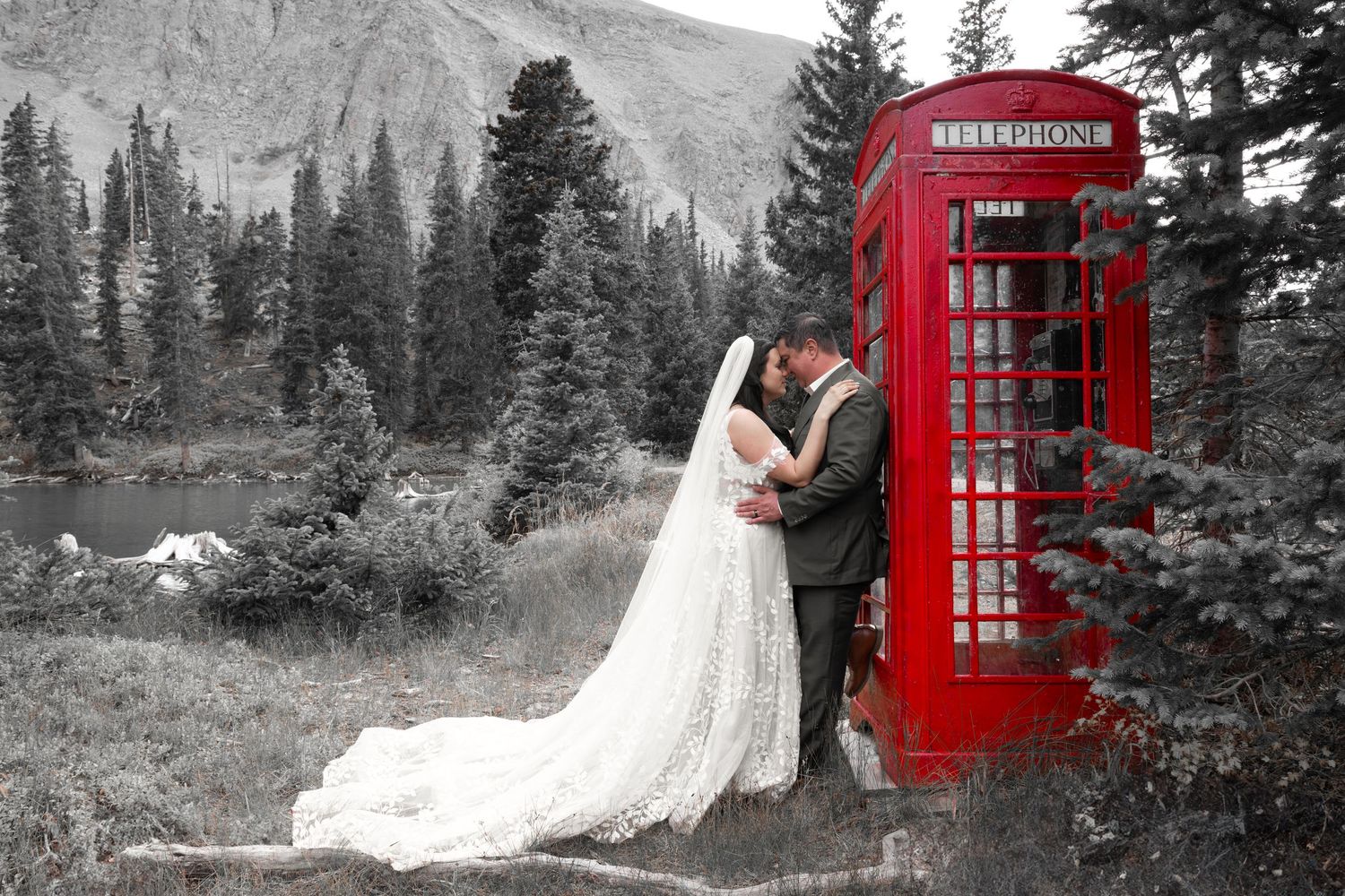 A bride and groom pose near a red British phone booth surrounded by pine trees in a mountain setting. The background is black and white.