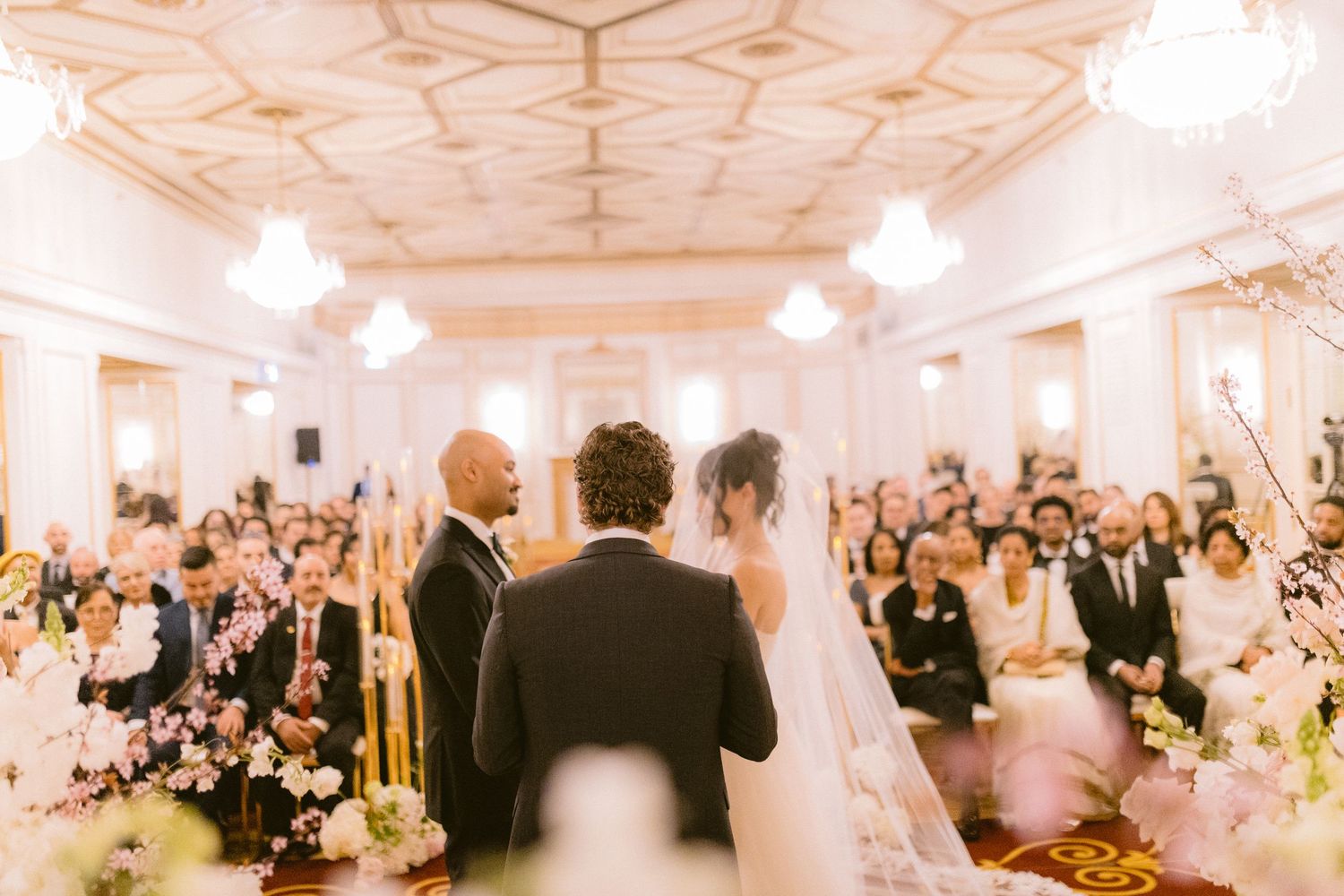 A dreamy wedding ceremony in an elegant white ballroom with ornate ceiling details and floral decorations.