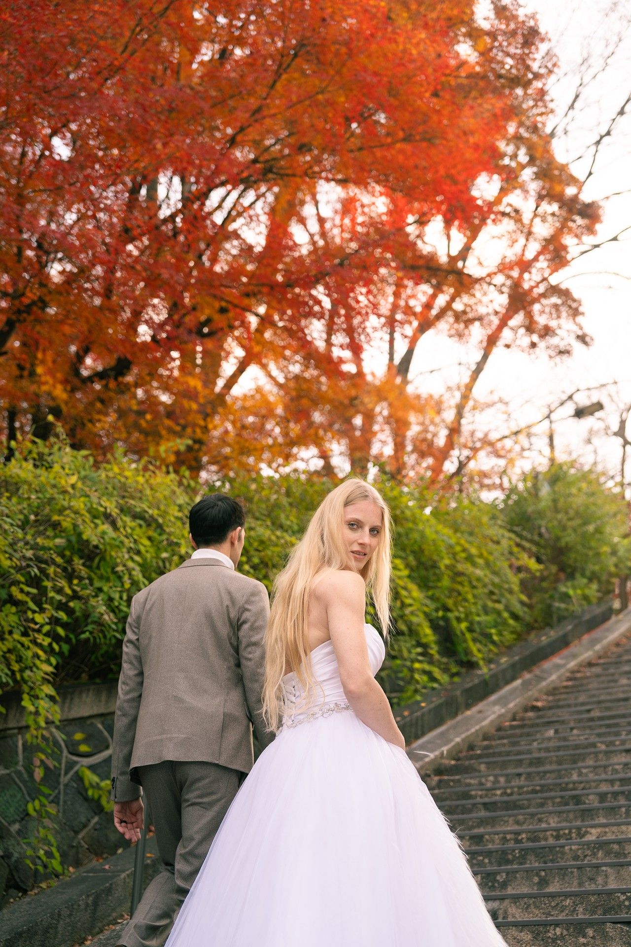 A couple in wedding attire ascend stone steps surrounded by autumn foliage and green hedges.