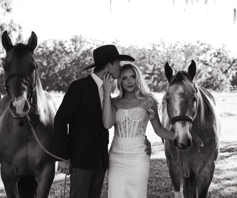 A romantic black and white photo of a couple in western attire standing between two horses in a rustic outdoor setting.