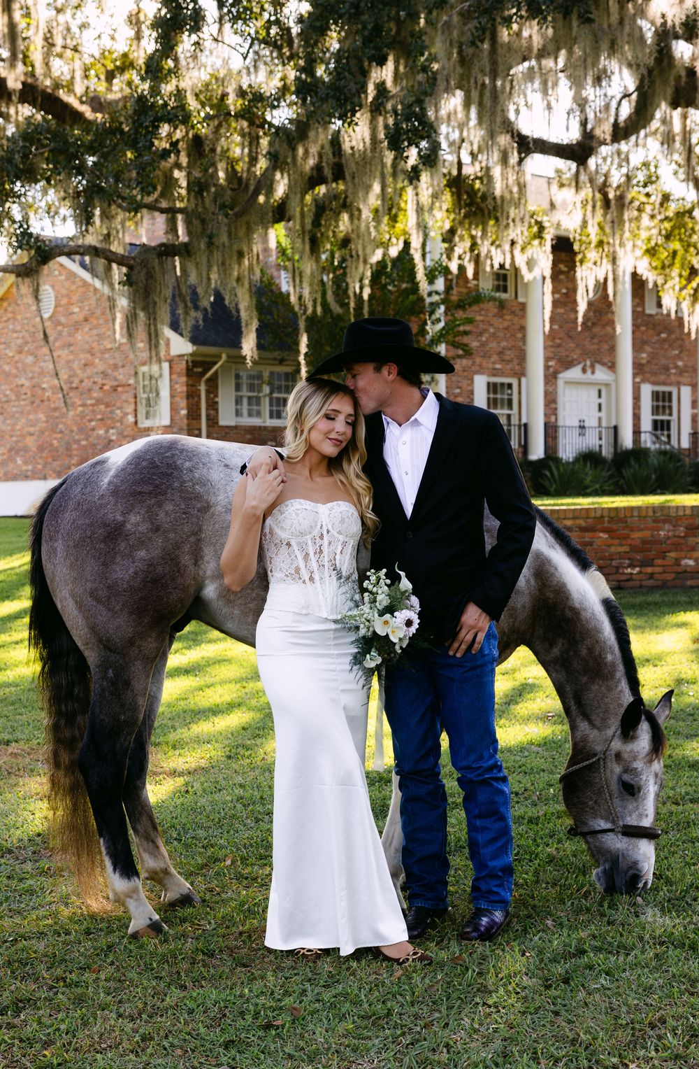 A romantic photoshoot in a southern garden with Spanish moss and a gray horse grazing beside a couple.