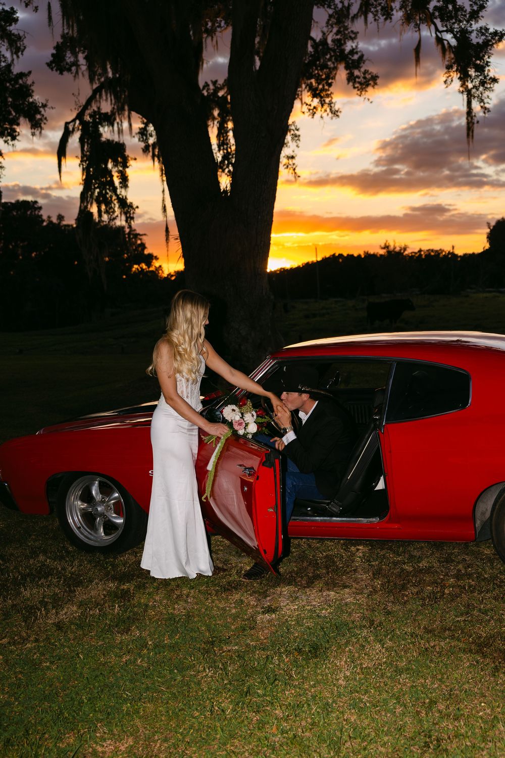 A sunset wedding photo with a classic red muscle car and dramatic orange sky.