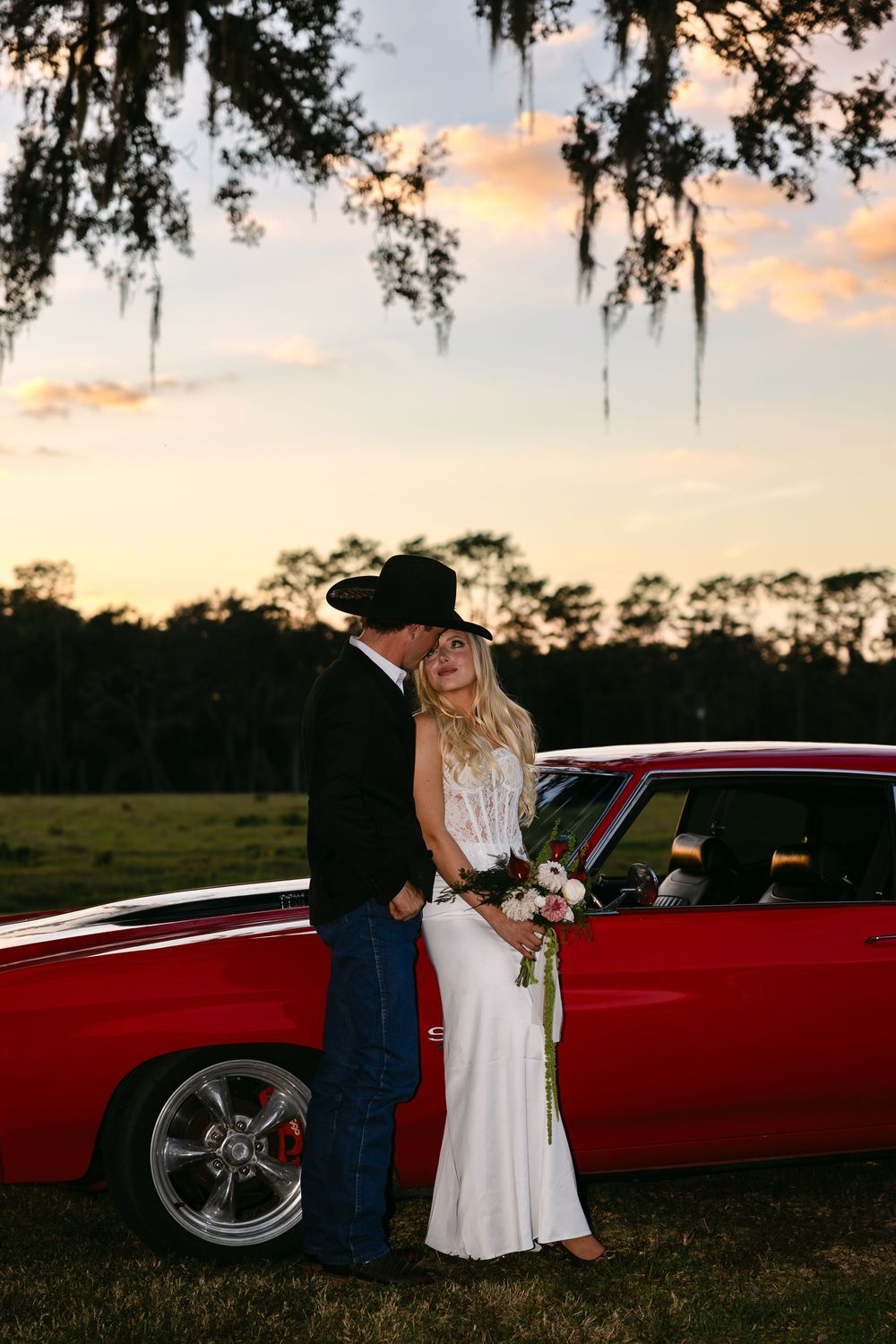 A couple poses at sunset next to a red classic car under Spanish moss-draped trees.