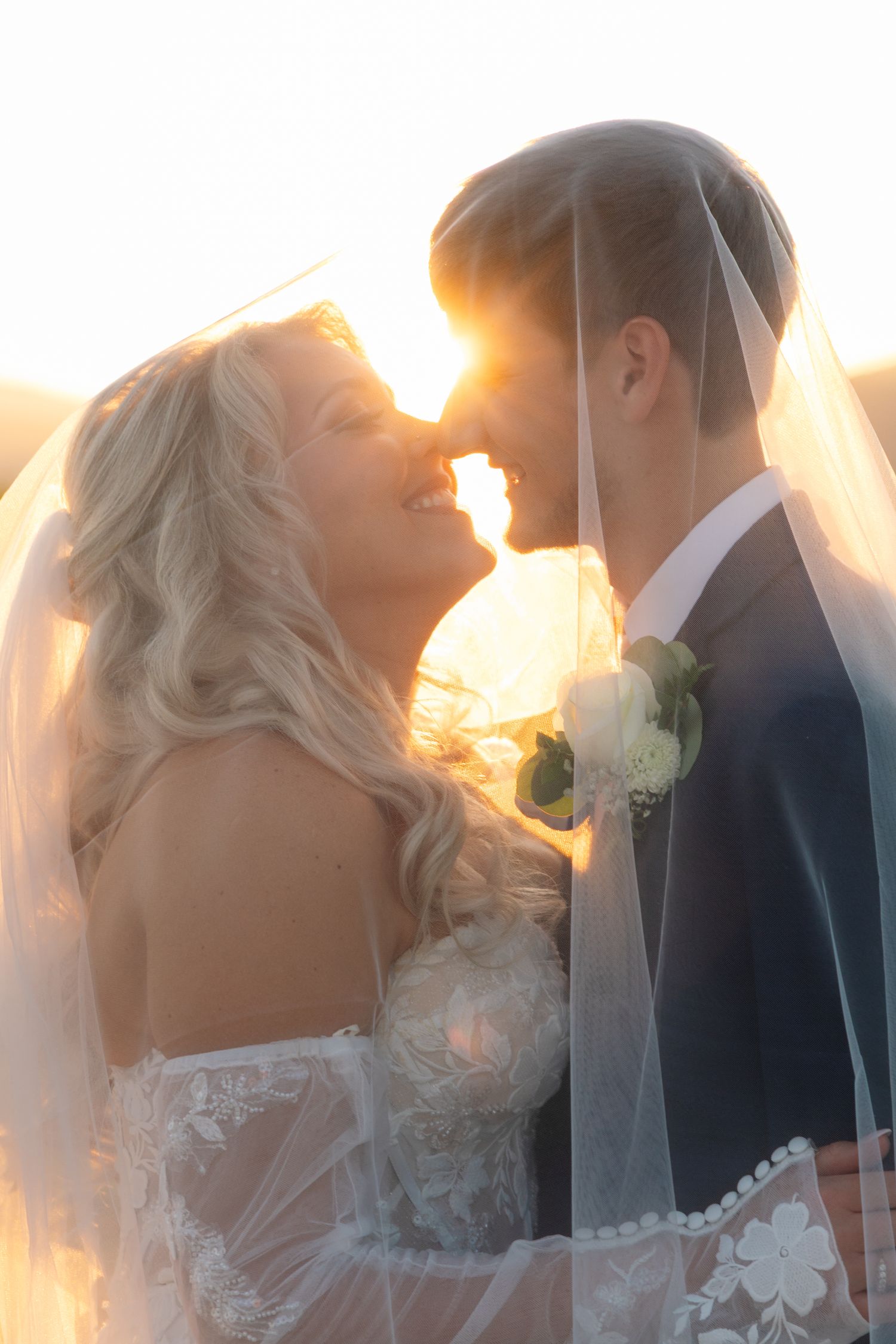 A romantic wedding couple shares an intimate kiss at sunset with a soft glowing backlight creating a dreamy atmosphere.