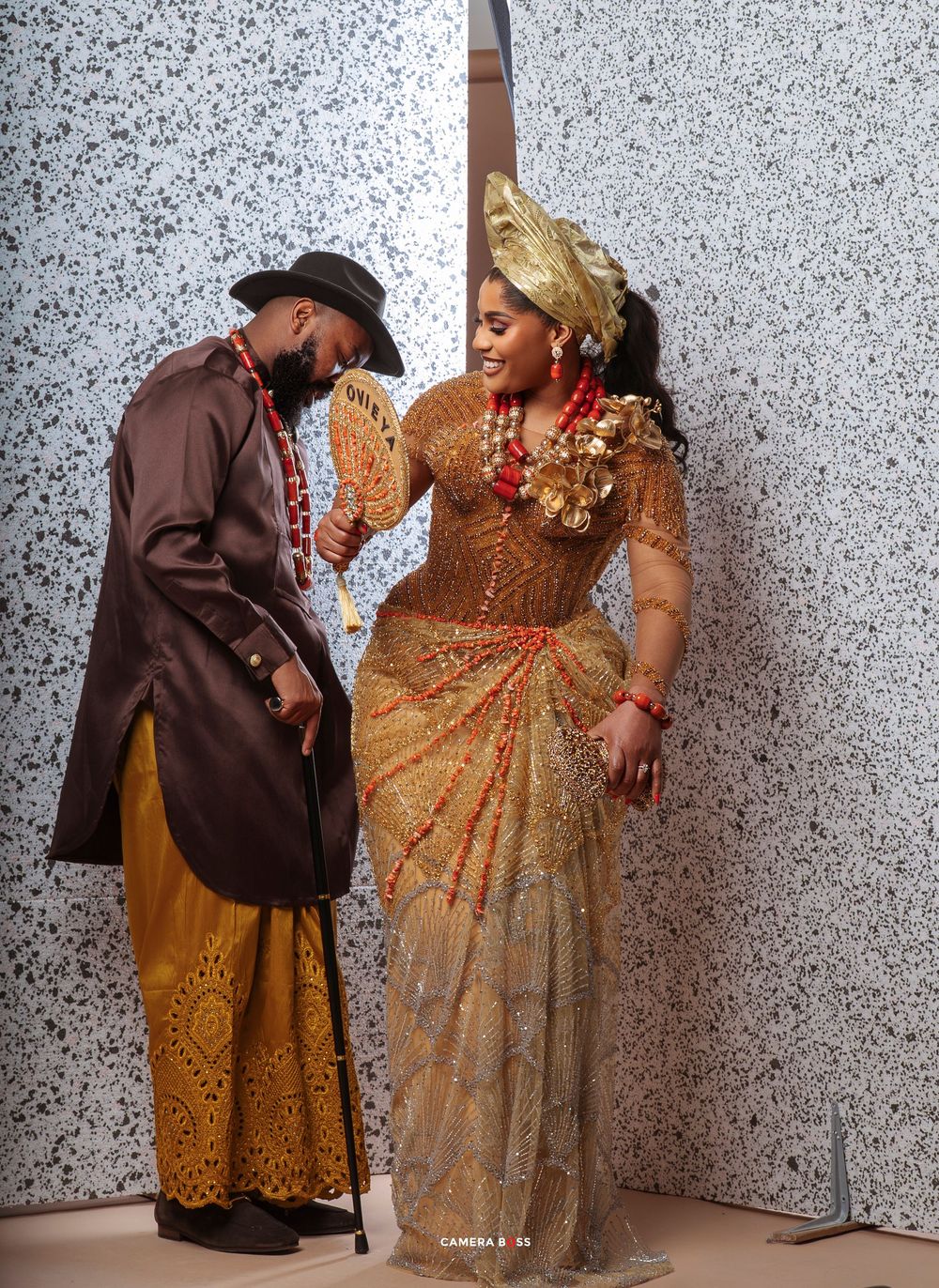 Two people in traditional African-inspired attire pose against a speckled wall, wearing elegant gold and brown outfits.